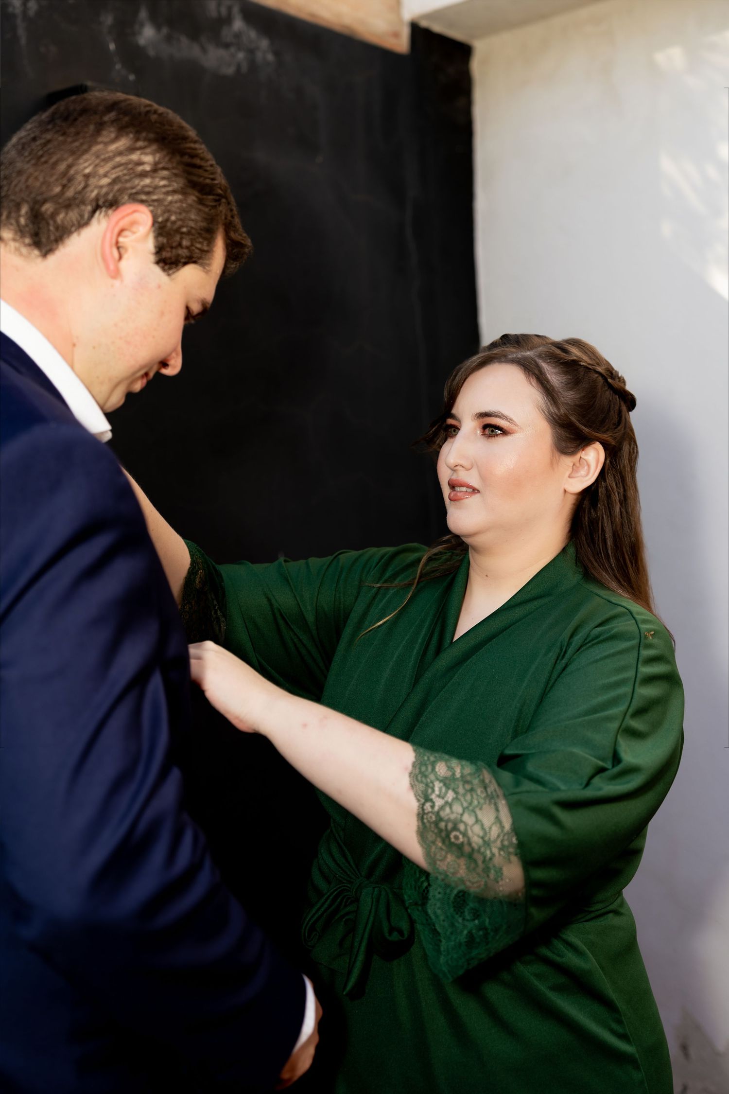Woman in green satin robe adjusting a man's navy suit jacket during wedding preparations.