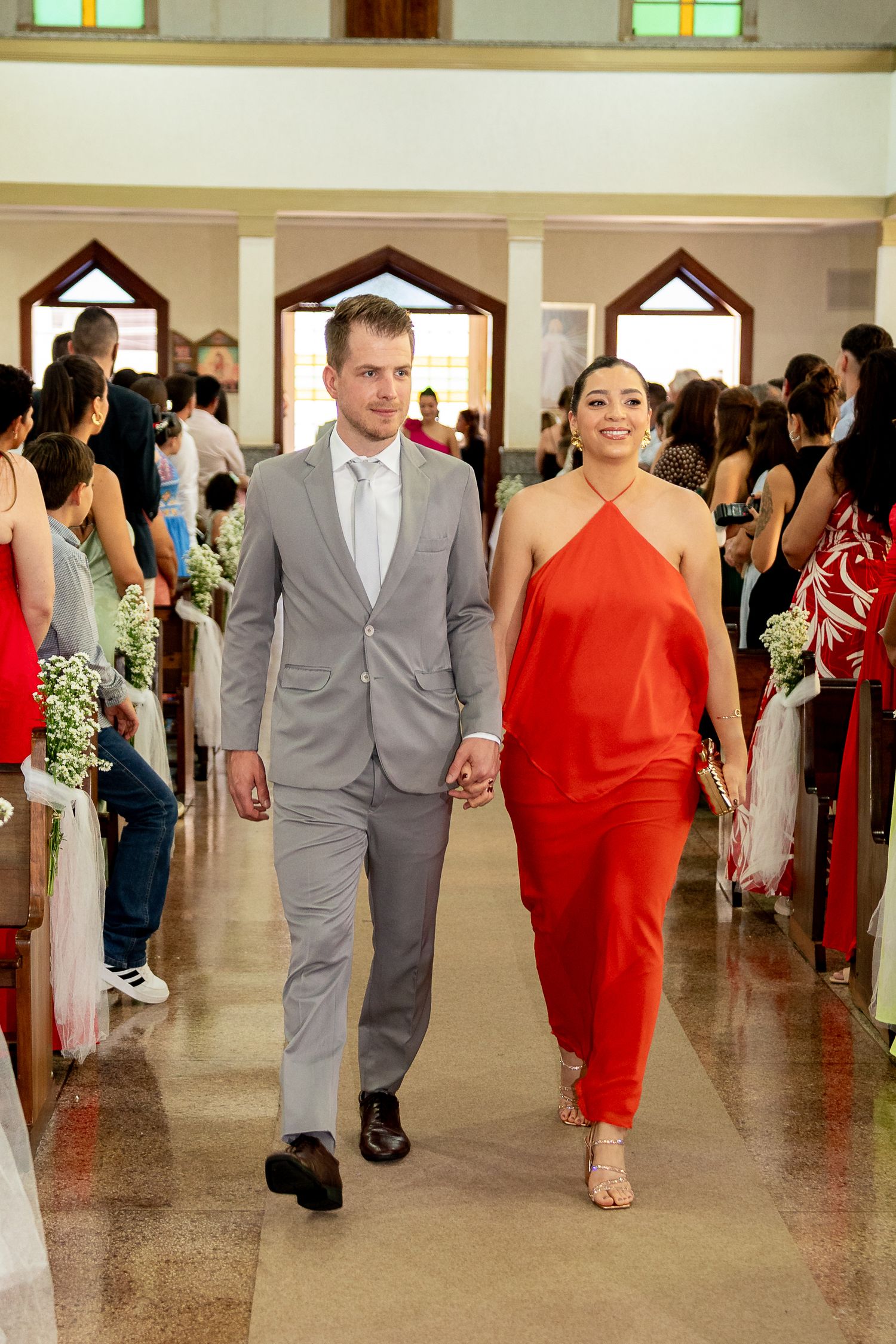 Couple walks down church aisle, man in gray suit, woman in red halter dress, guests seated on both sides.