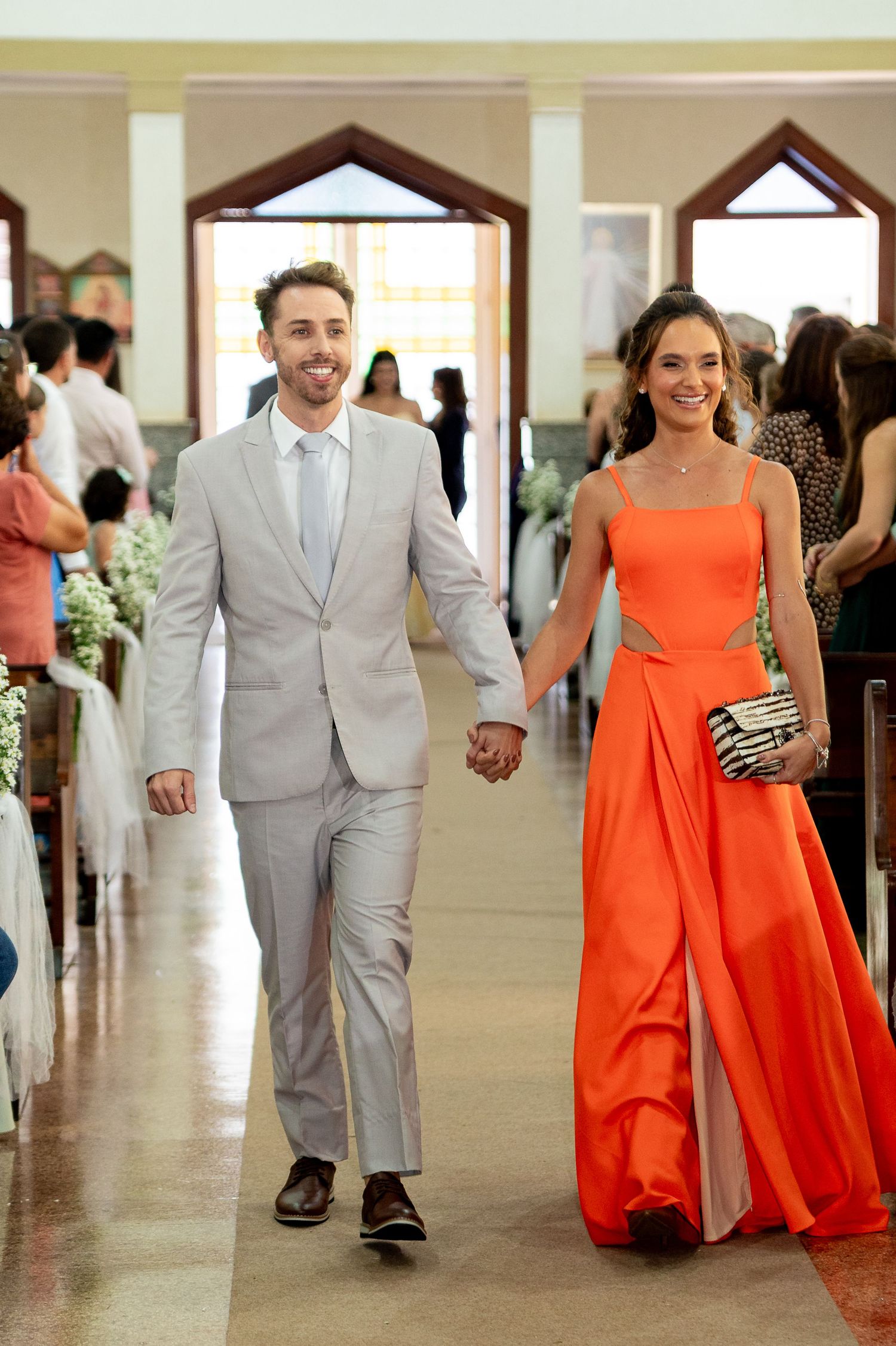 Couple walking down church aisle, man in gray suit, woman in elegant orange gown, holding hands at wedding.