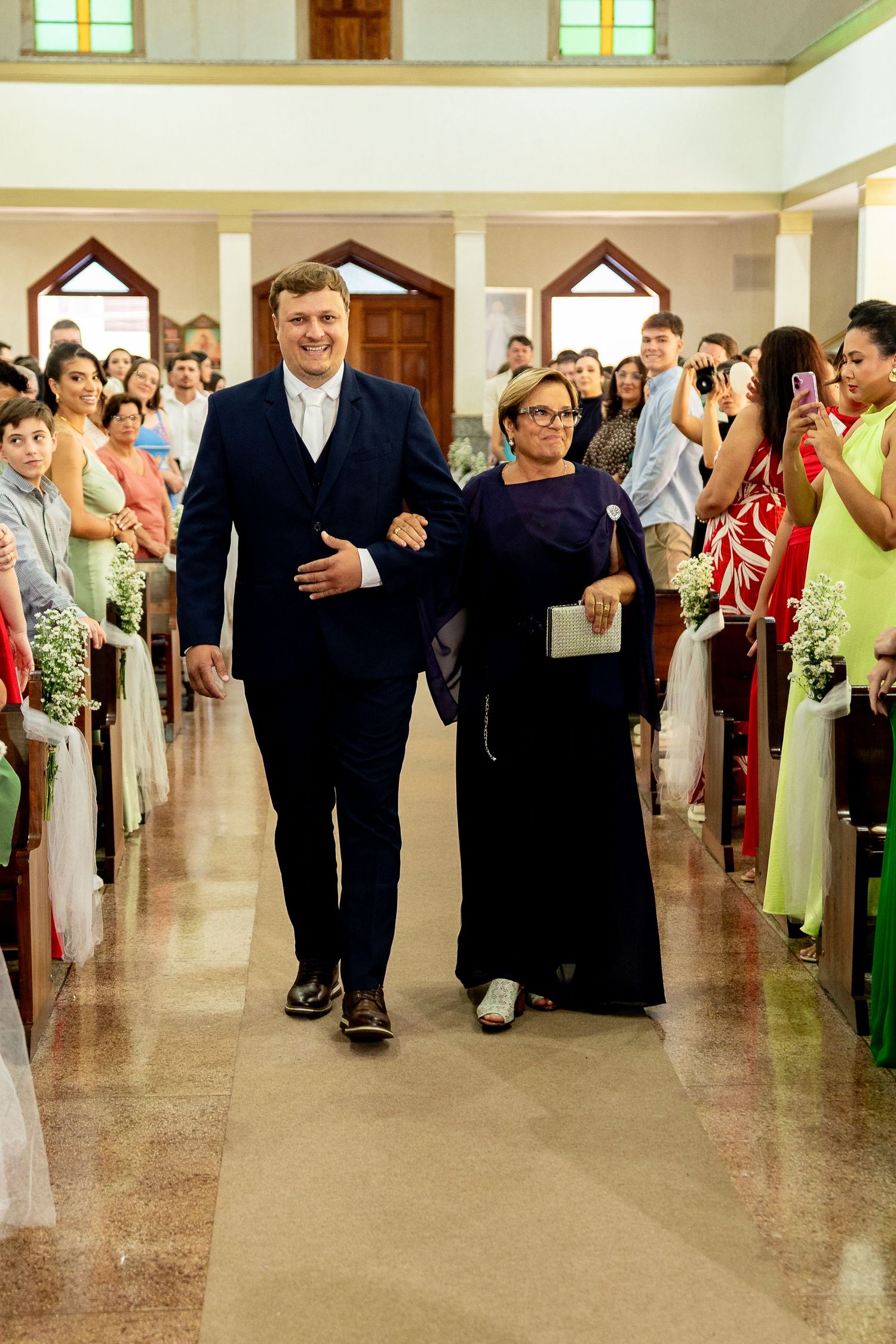 Groom in navy suit walks down church aisle with older woman during wedding ceremony, guests watching.