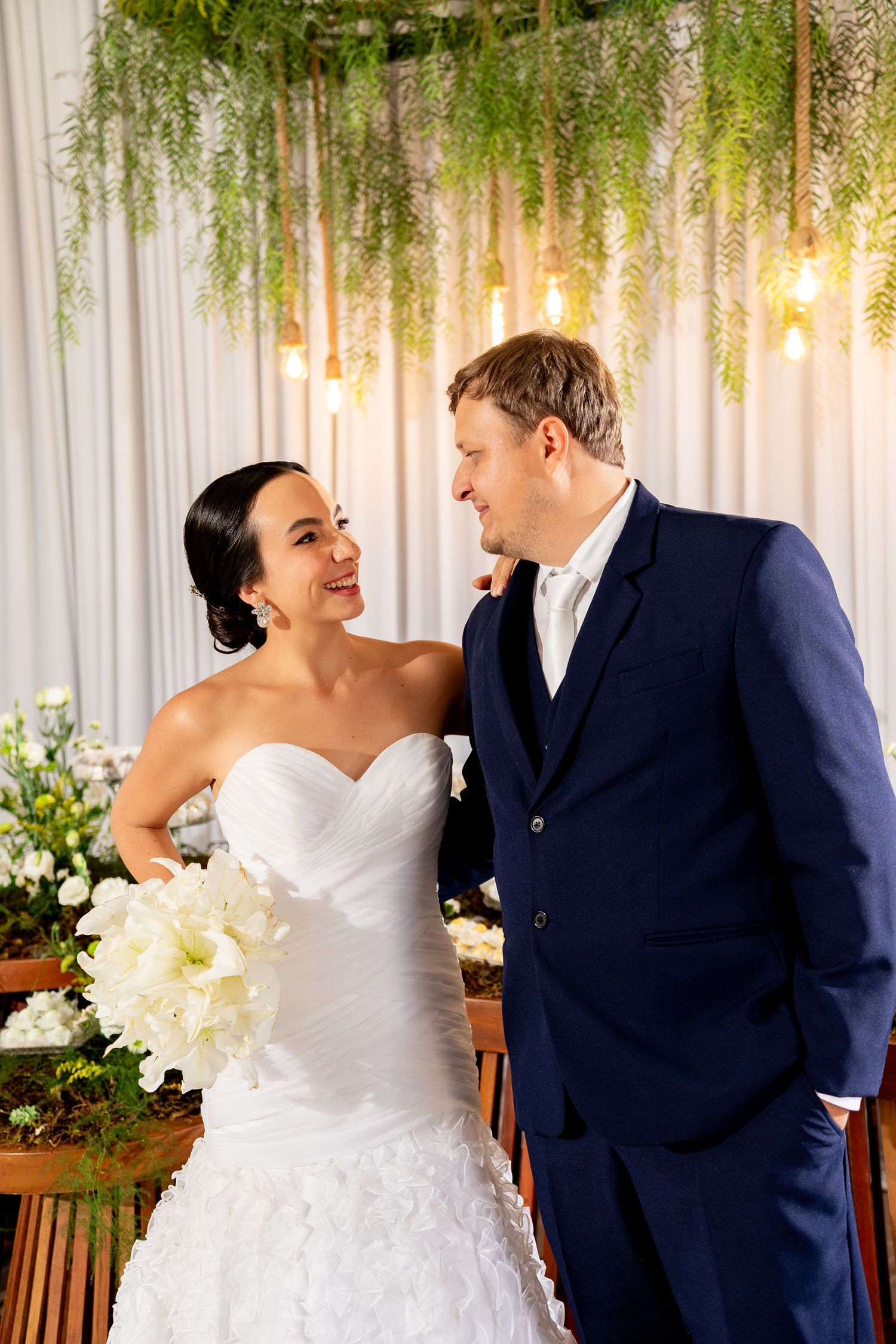Bride in white strapless gown holding white bouquet smiles at groom in navy suit at elegant wedding ceremony.