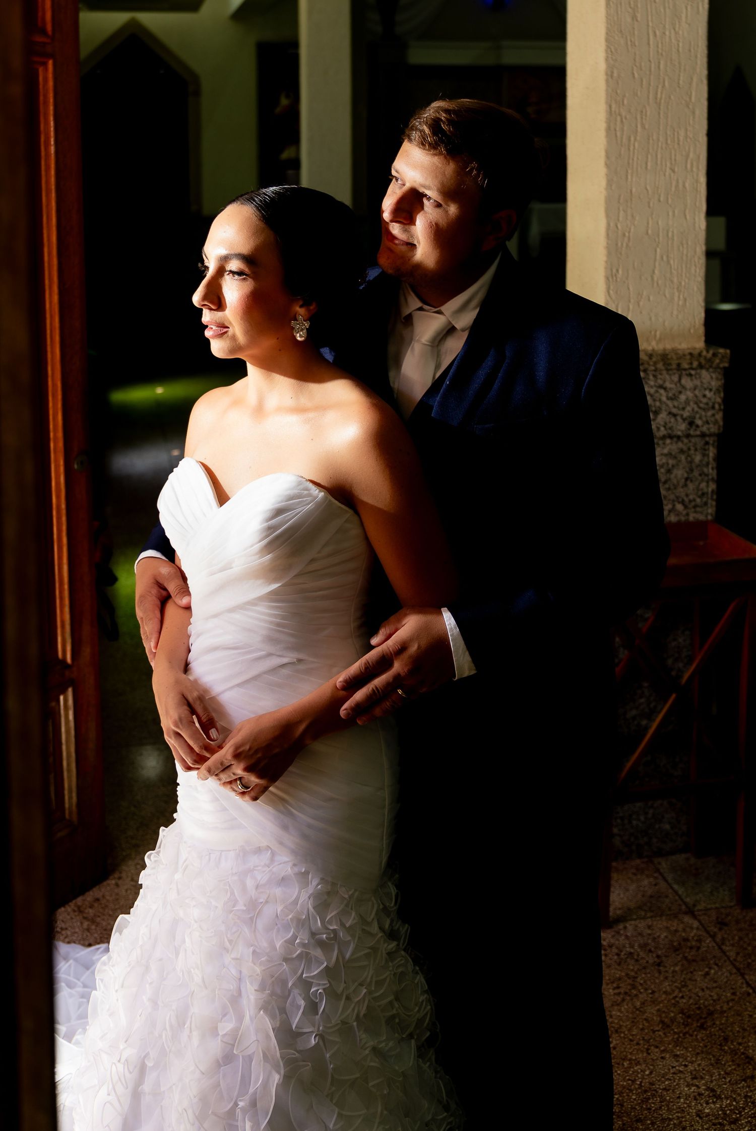 Wedding couple posing romantically in doorway, bride in strapless ruffled gown, groom in navy suit.