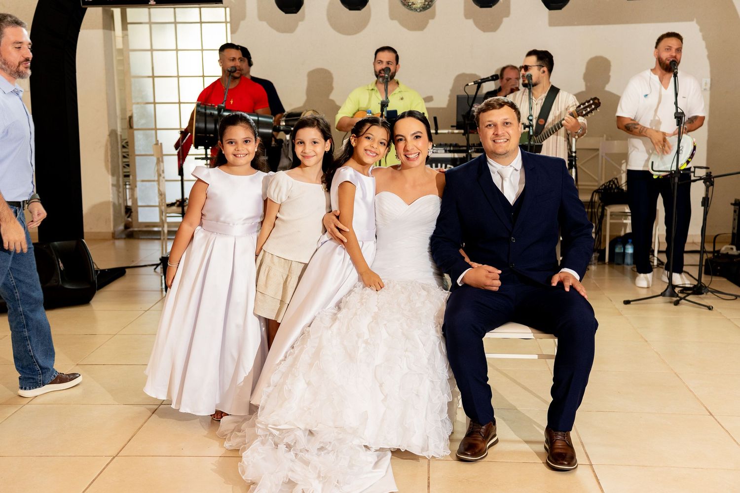 Bride and groom pose with two flower girls on a white dance floor during a wedding reception with live band.