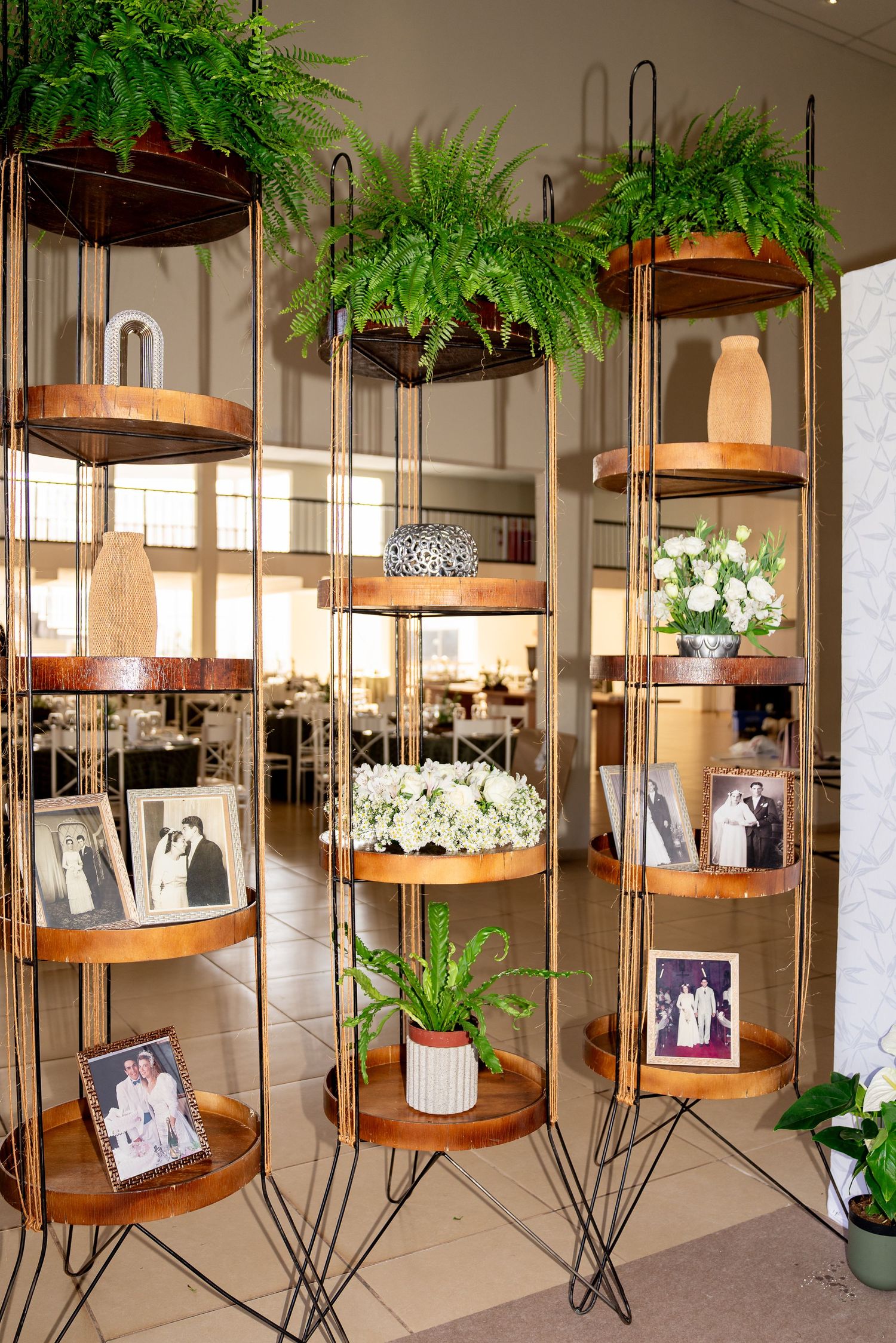 Elegant tiered metal shelving units with copper trays display ferns, white flowers, and vintage wedding photos at an event.