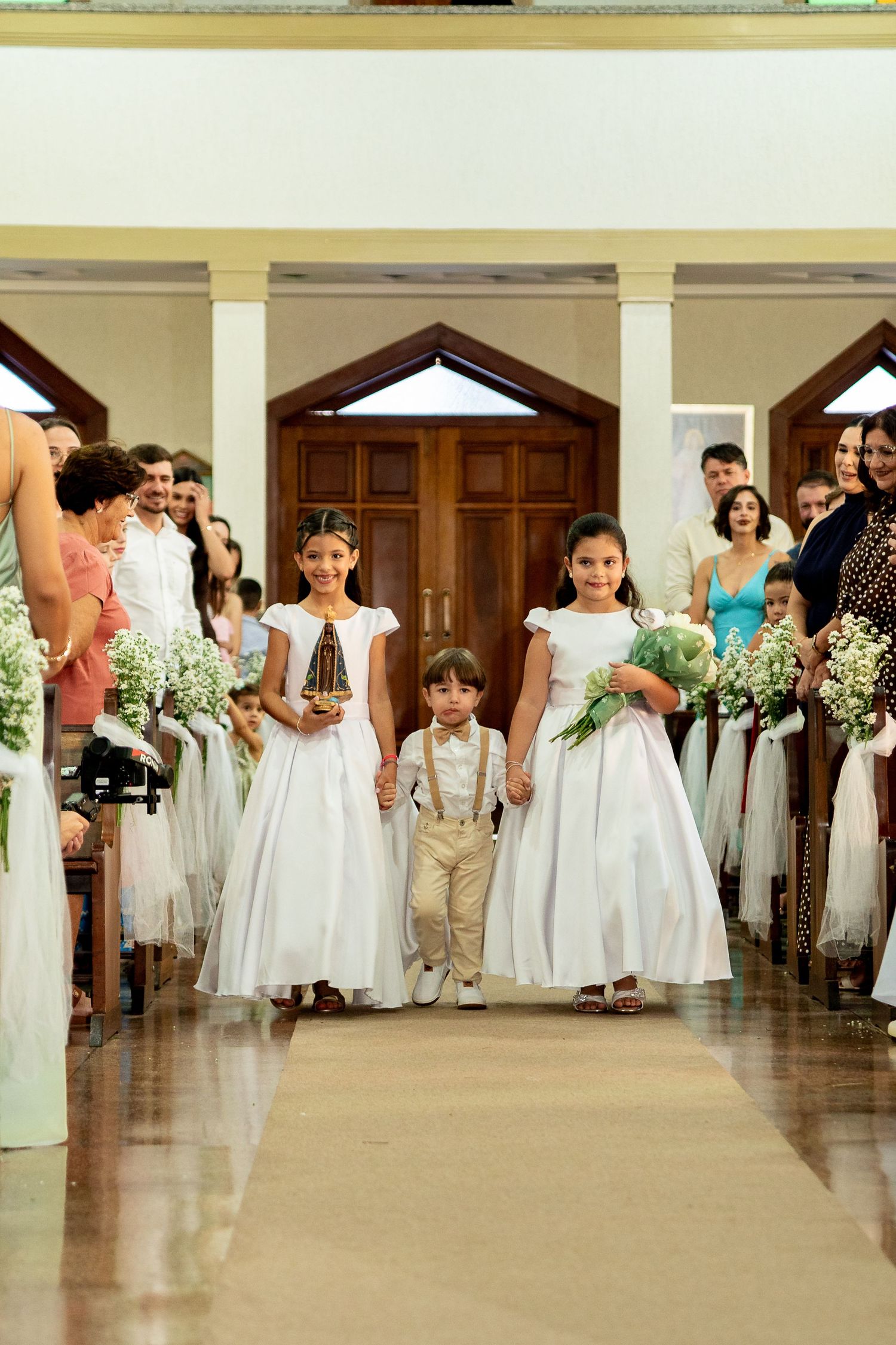 Two flower girls in white dresses and a ring bearer walk down the wedding aisle in a church ceremony.