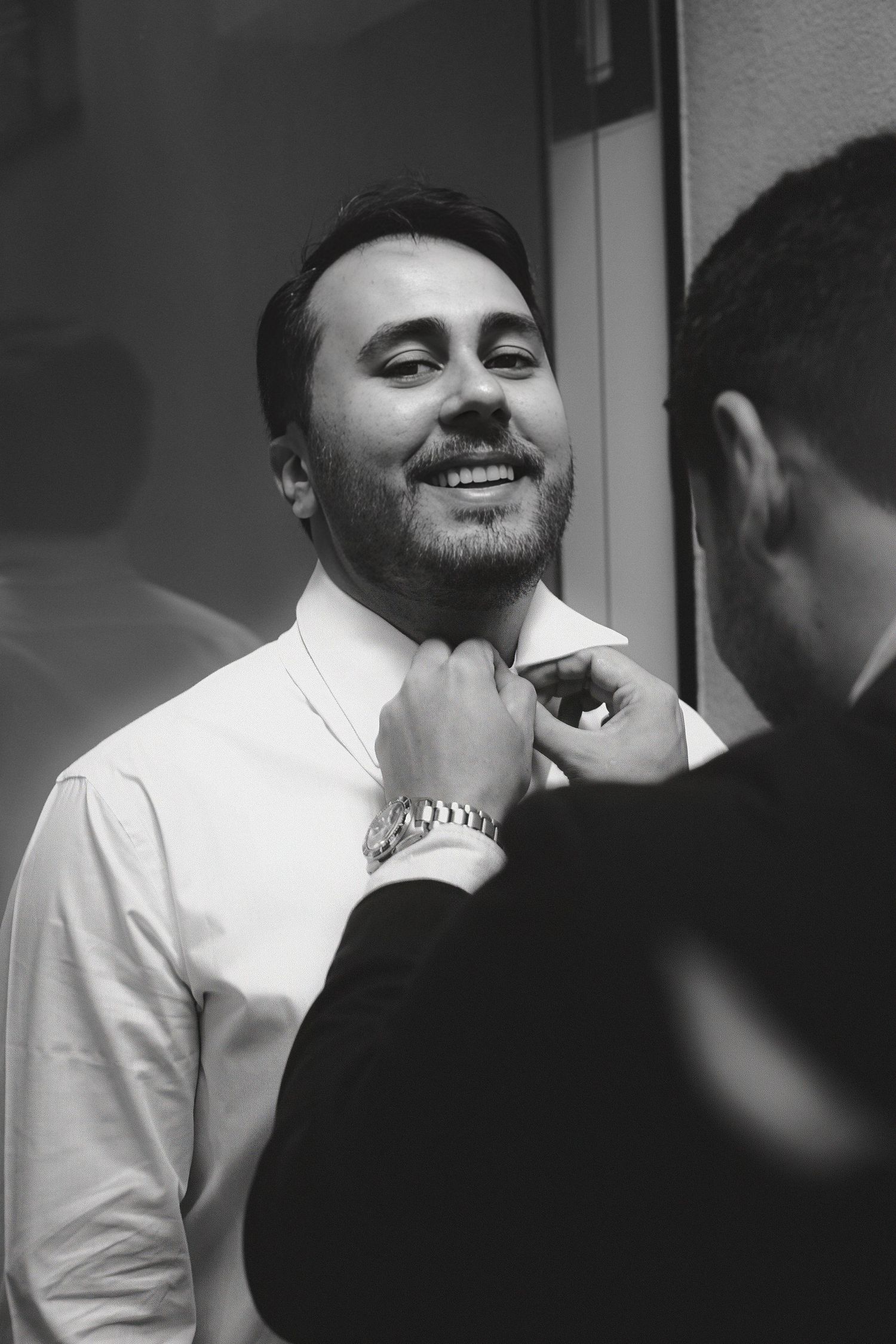 Black and white photo of a smiling groom having his tie adjusted by a groomsman before the wedding ceremony.