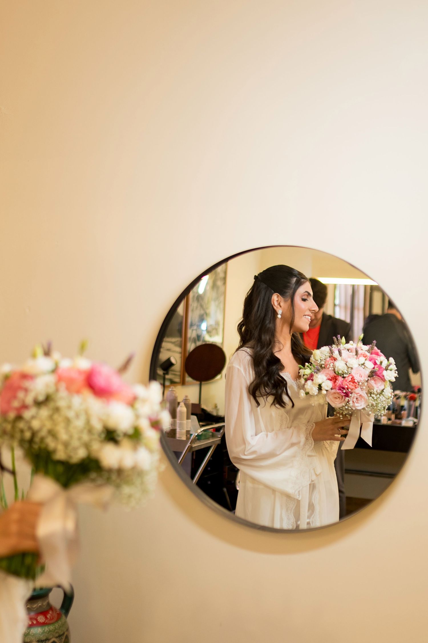 Bride in white robe holding pink bouquet reflected in round mirror during wedding preparations.