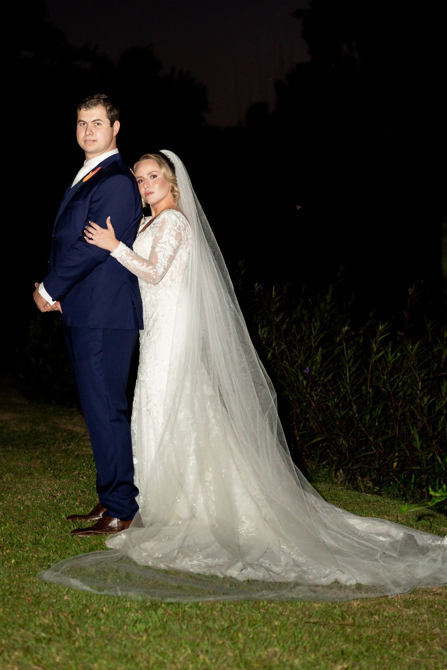Bride in lace gown with veil and groom in navy suit pose back-to-back outdoors at night.