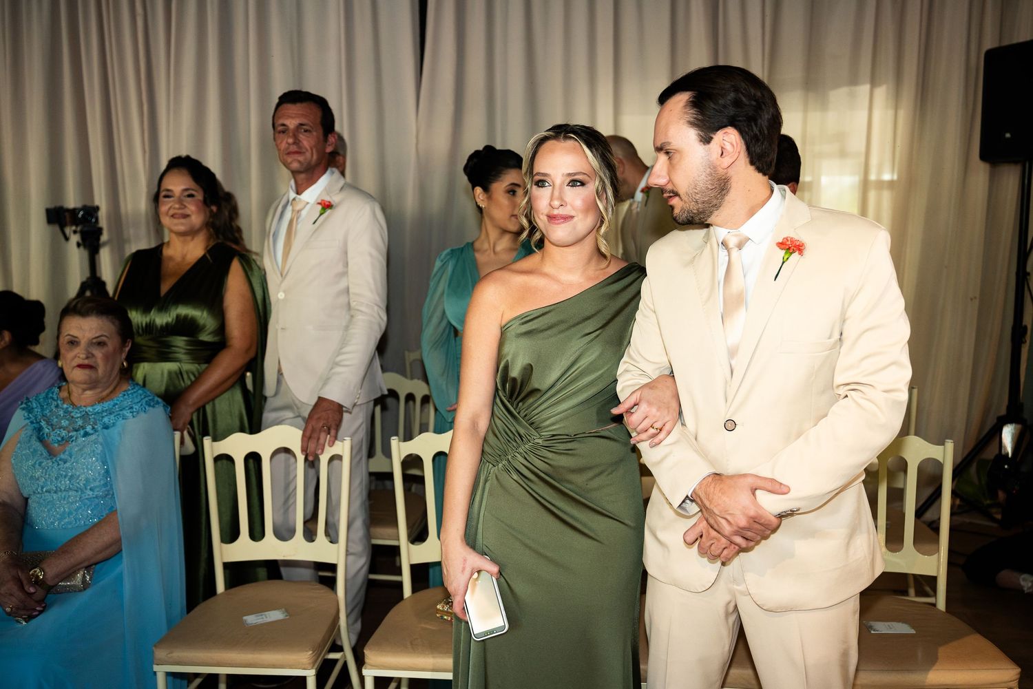 Elegant wedding guests in olive green and cream attire standing in a decorated venue during a ceremony.