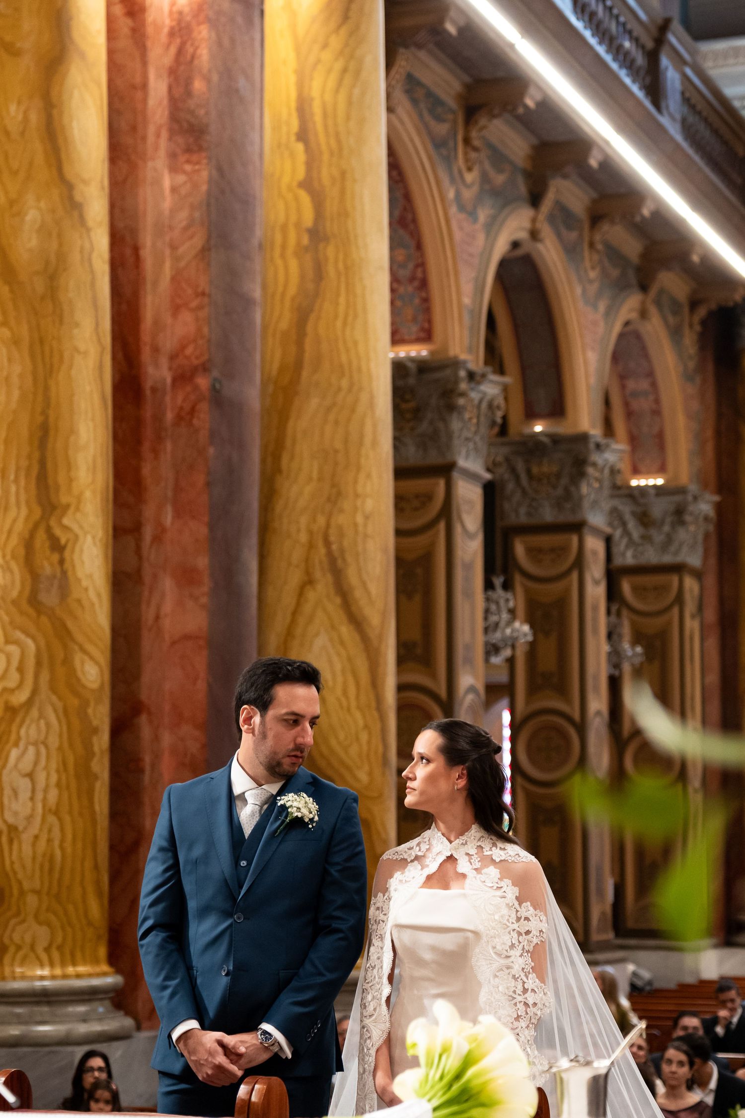 Bride in lace cape veil and groom in blue suit stand at church altar during elegant wedding ceremony.