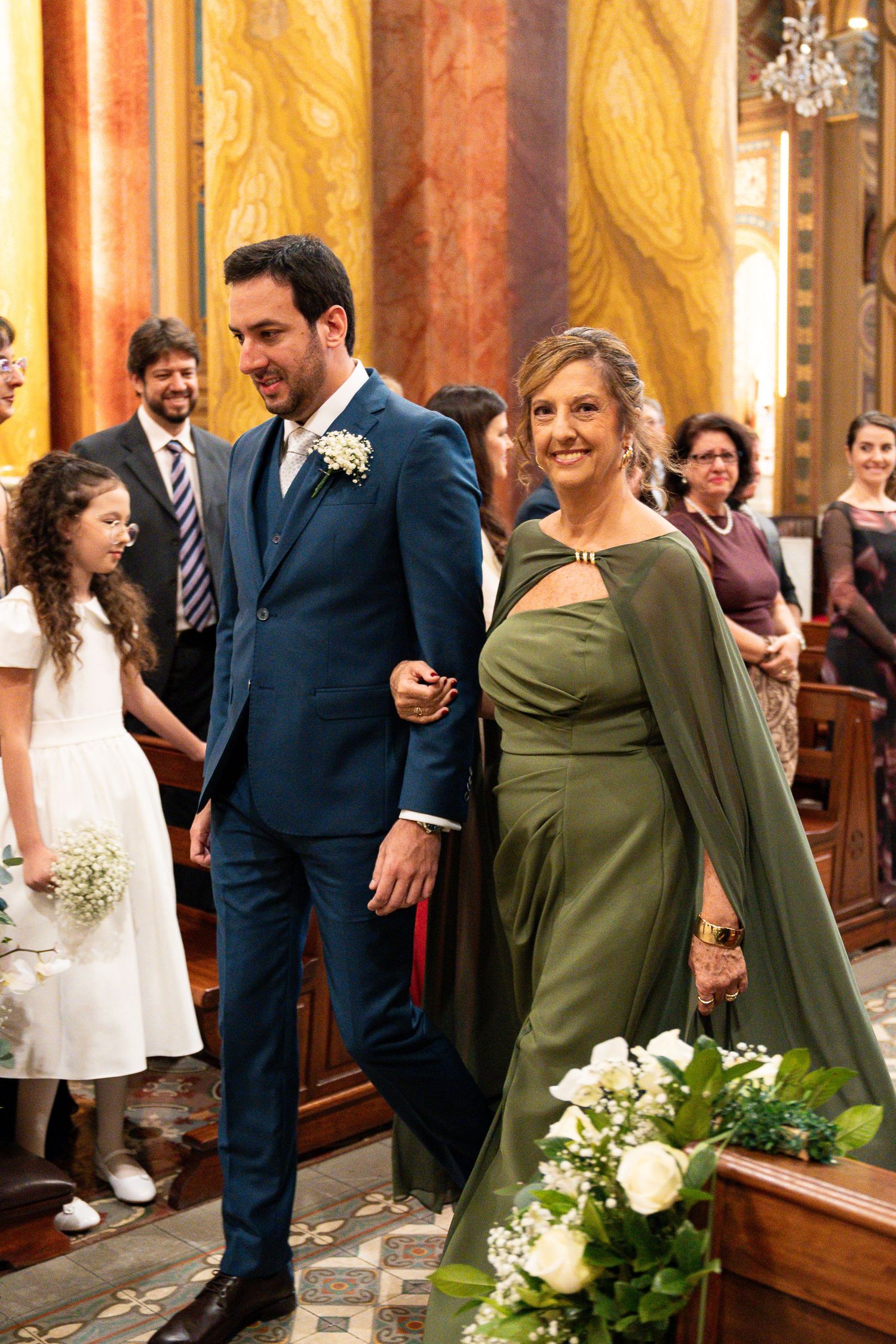 Groom in blue suit walks with woman in green gown down church aisle at elegant wedding ceremony.