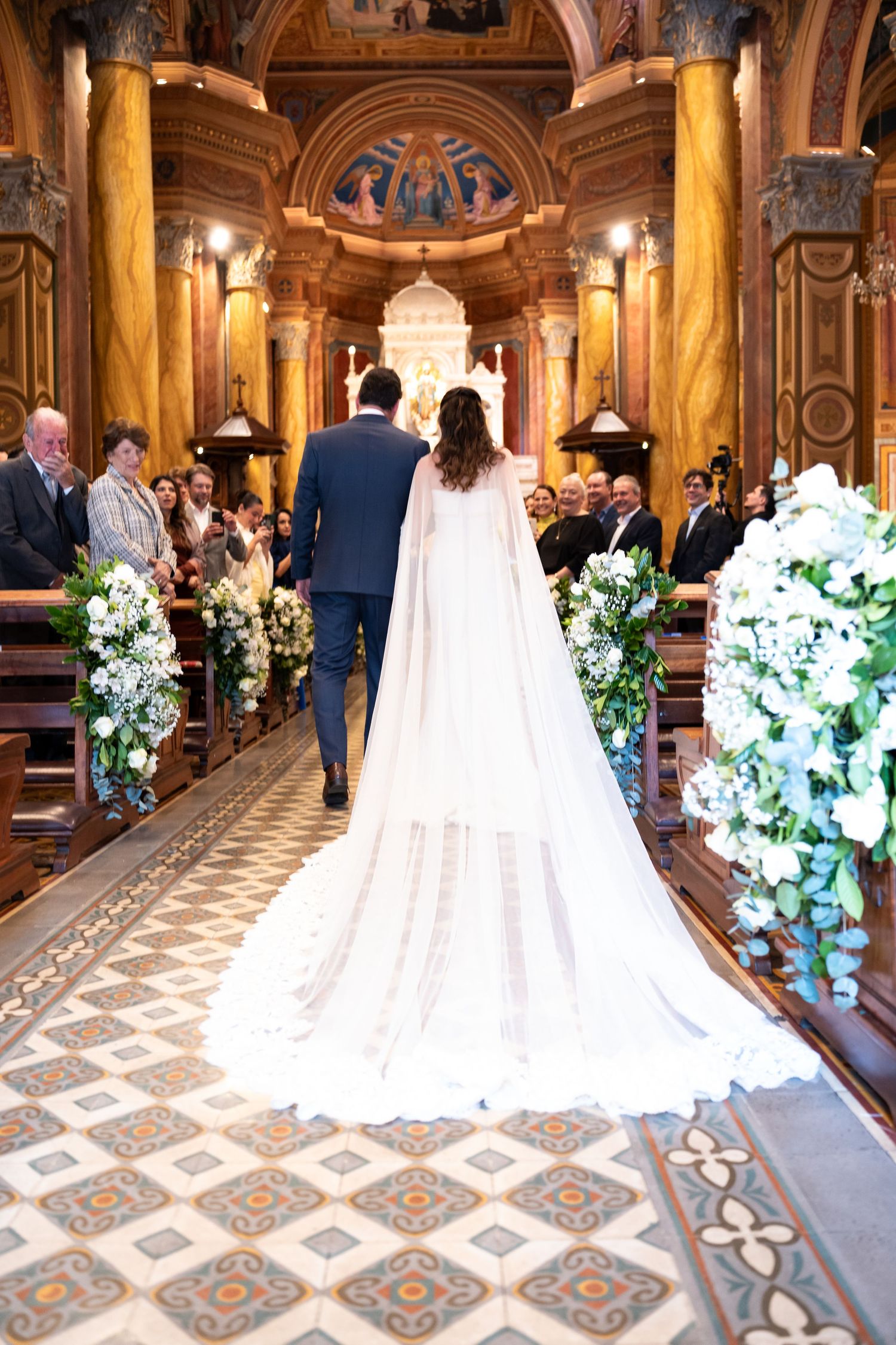 Bride and groom walking down the aisle of an ornate church, surrounded by white floral arrangements and wedding guests.