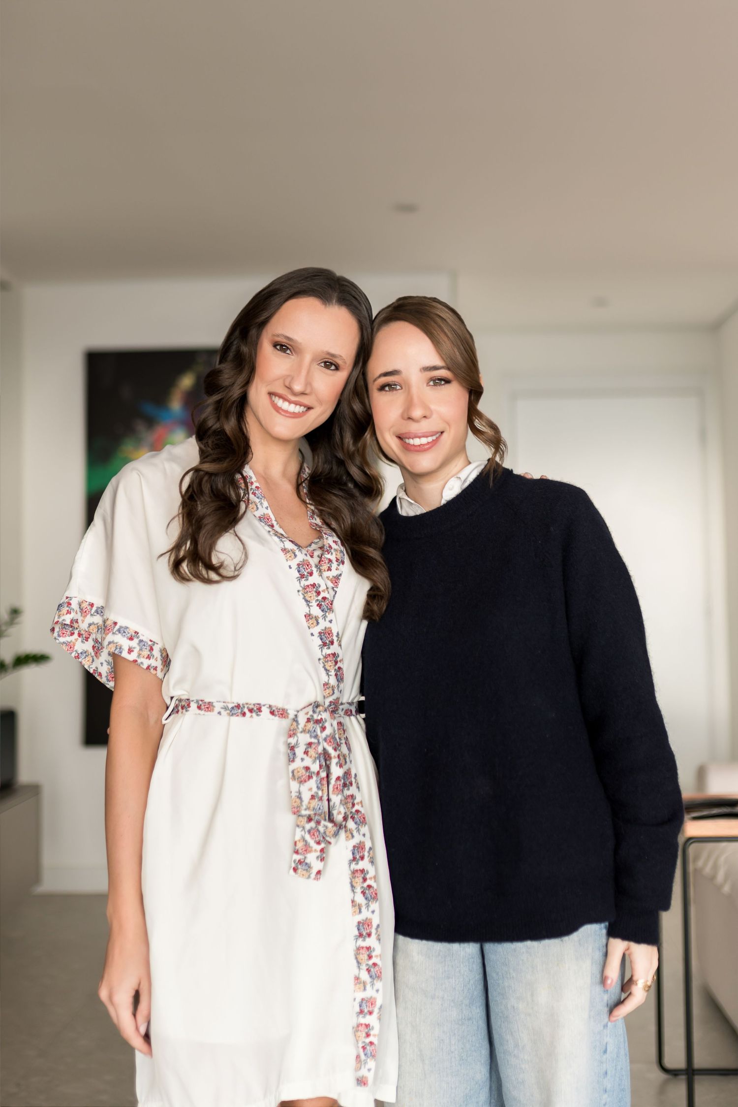 Two smiling women pose together indoors, one in a white floral dress and one in a navy sweater with jeans.