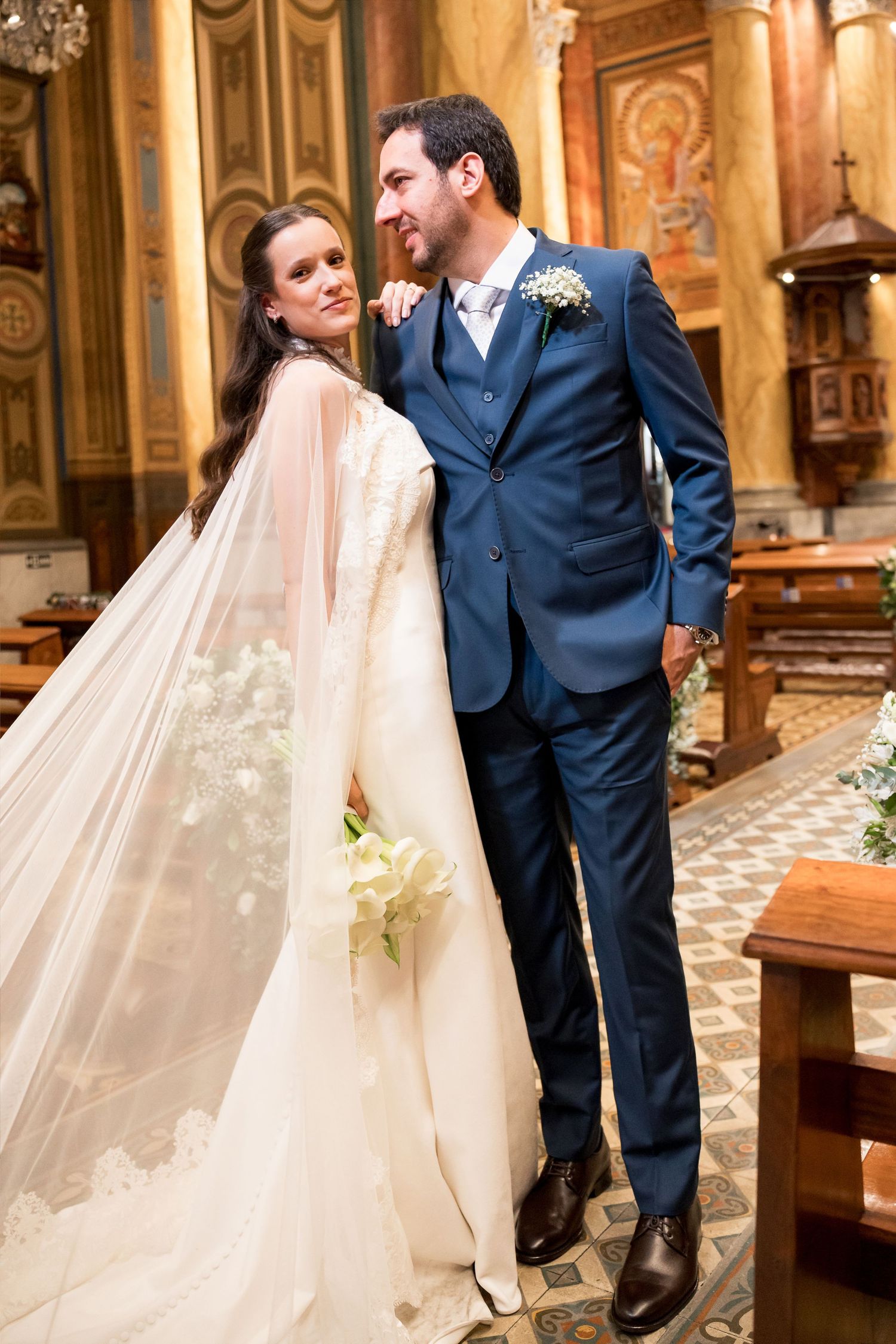 Bride in white gown and veil holds bouquet beside groom in blue suit inside ornate church.