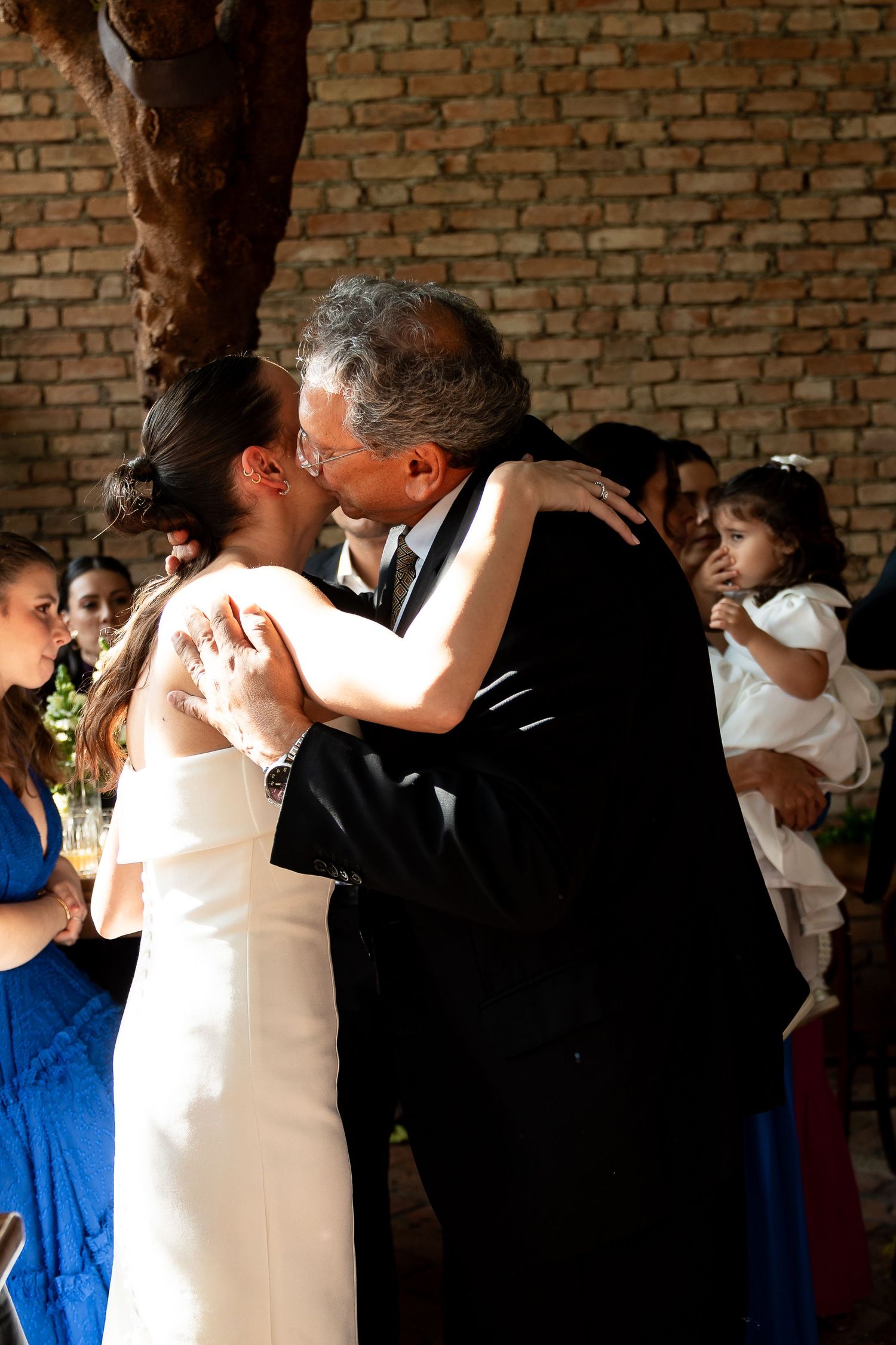 Bride and groom share their first kiss at wedding ceremony, surrounded by guests in a brick-walled venue.
