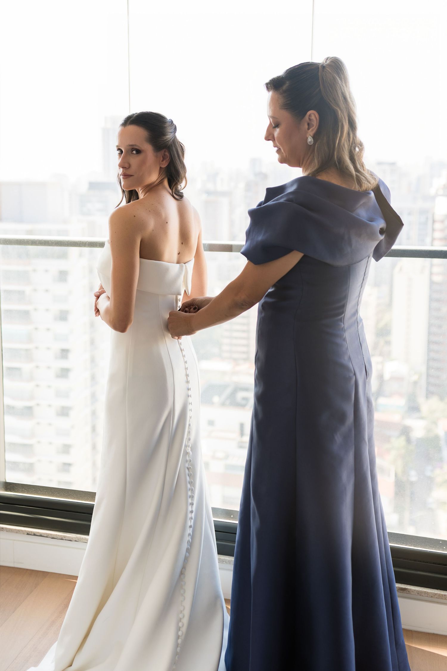 A bride in a white strapless gown gets her dress fastened by a woman in a navy blue off-shoulder dress near a window.
