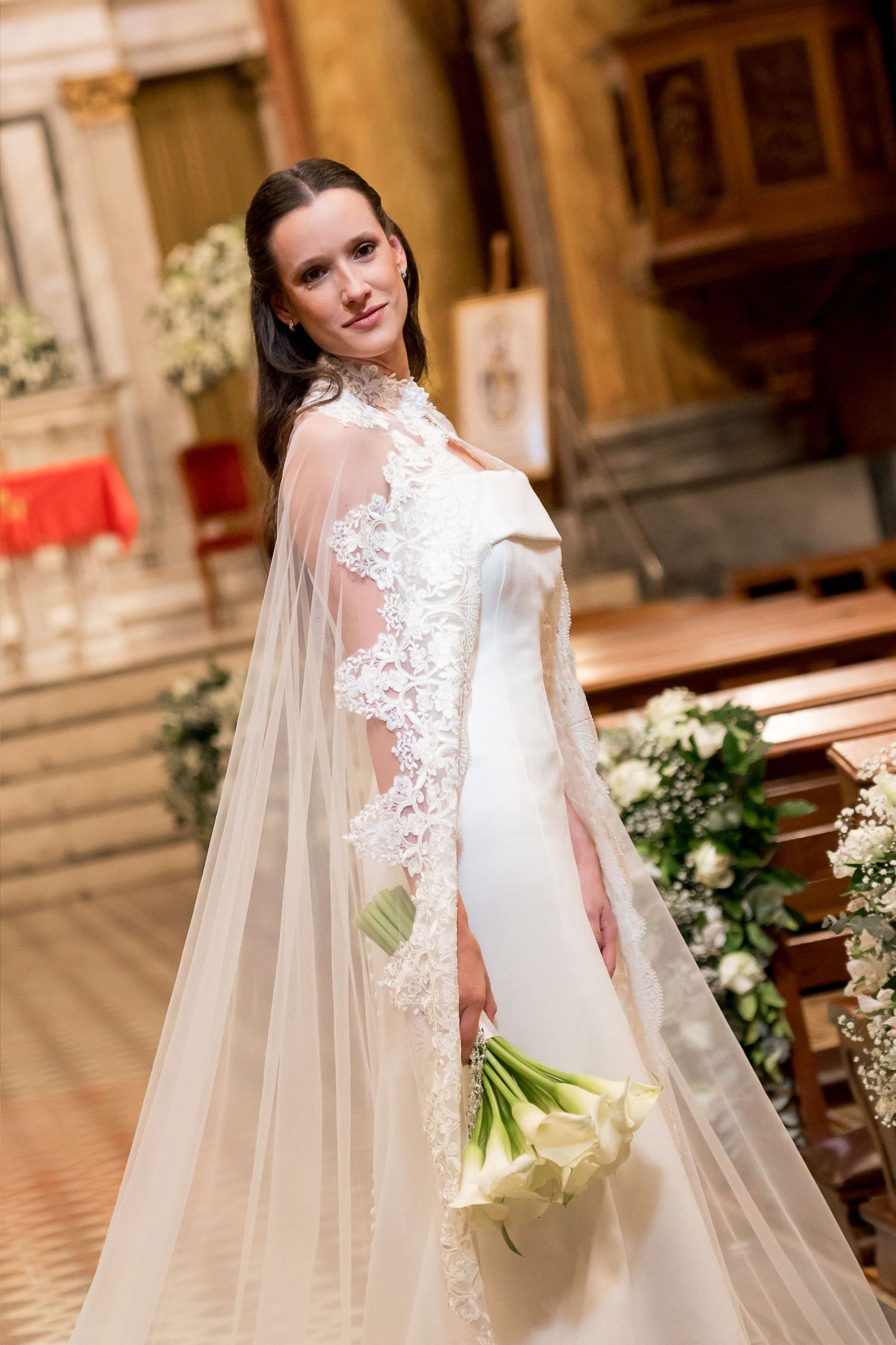Elegant bride in white lace cape wedding gown holding calla lily bouquet inside a beautiful church.