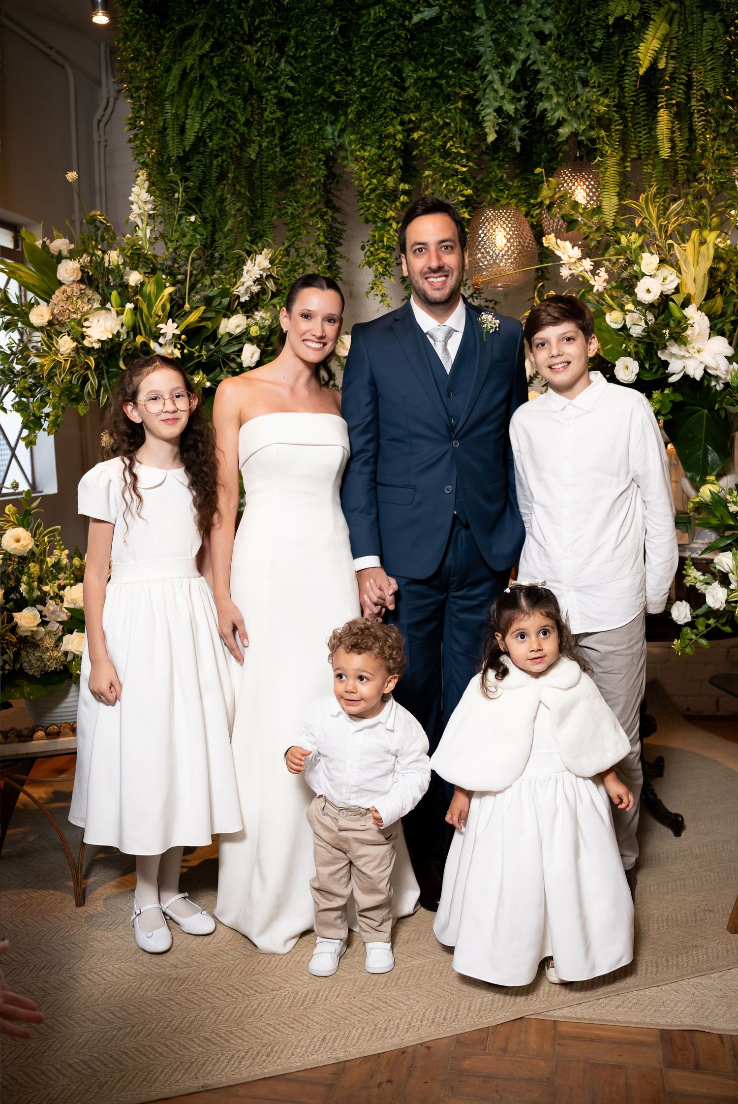Bride and groom pose with four children at elegant wedding ceremony with lush greenery and white floral backdrop.