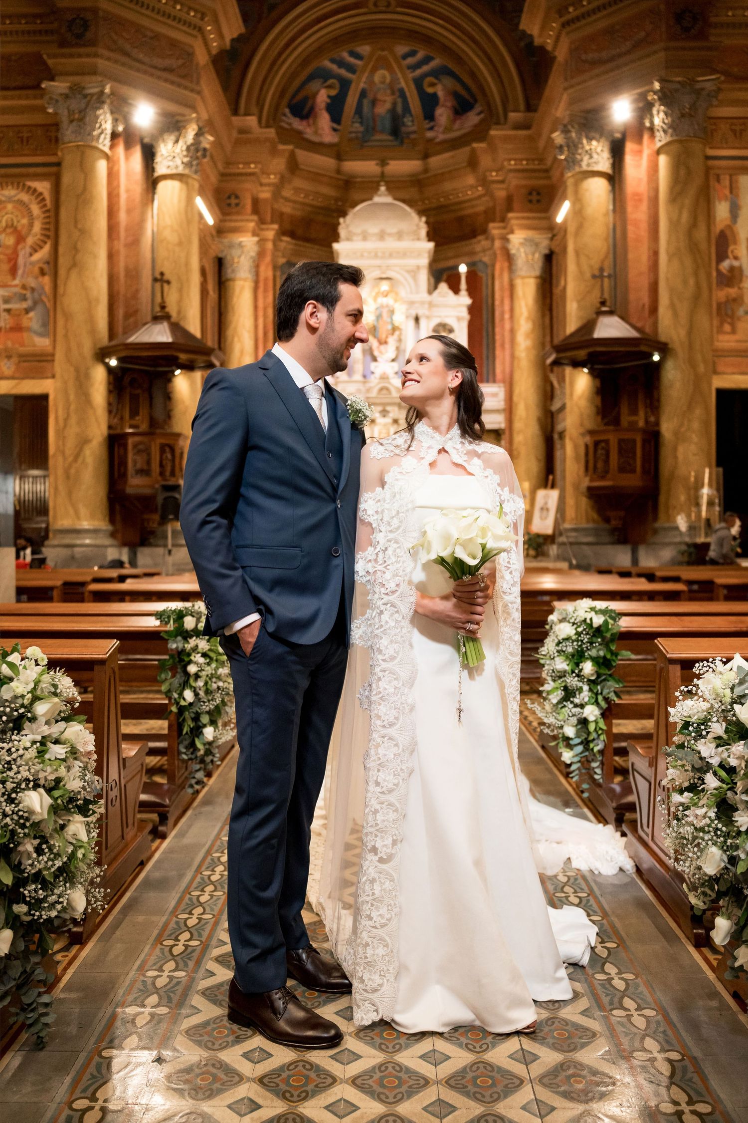 Bride and groom share a romantic moment at the church altar, she holds white roses bouquet wearing lace wedding gown.
