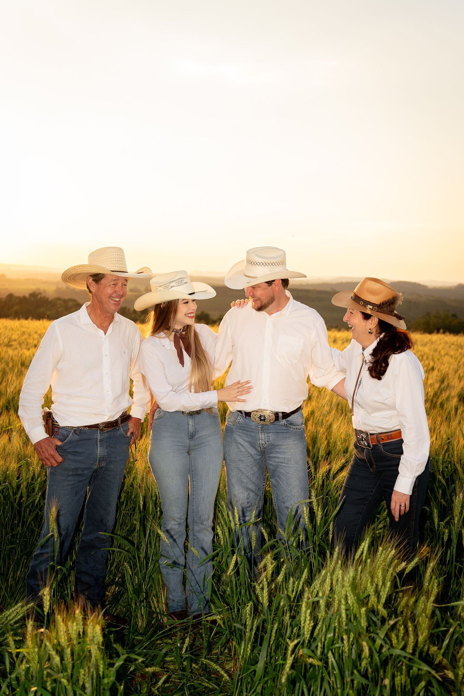 Four people in white shirts, jeans, and cowboy hats stand in a golden wheat field at sunset, smiling and talking.