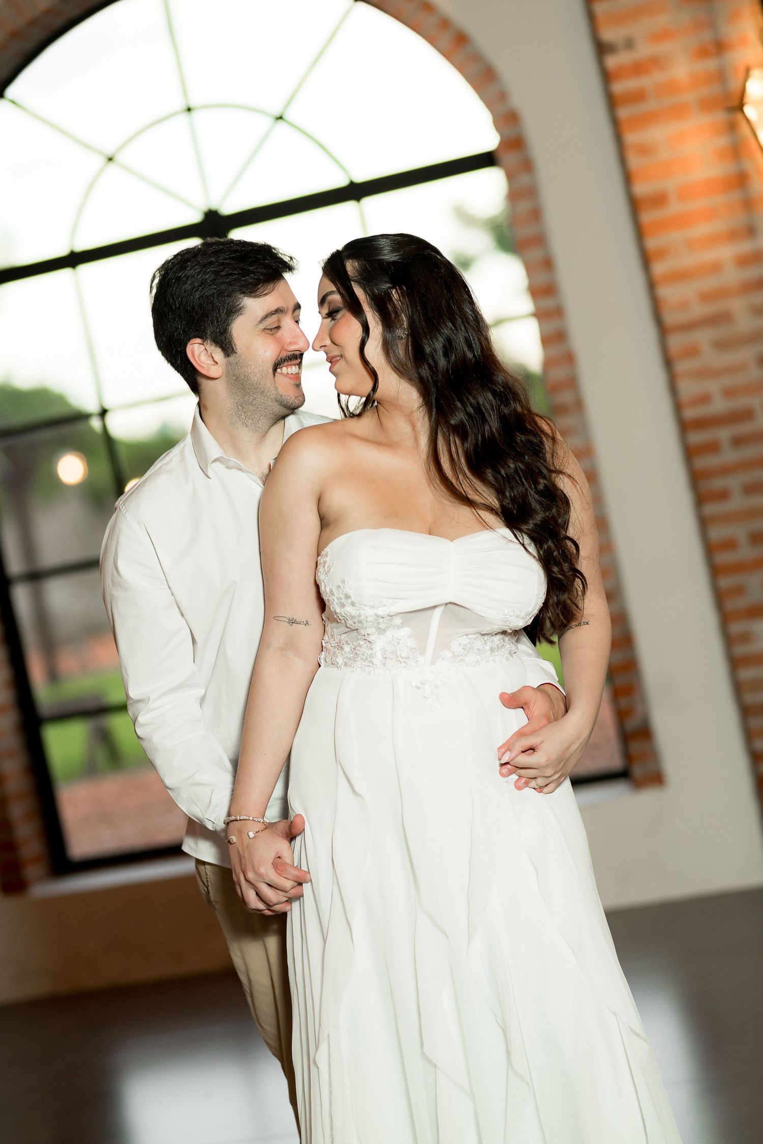 A couple in white wedding attire share a romantic moment, smiling at each other in an elegant brick-walled venue.