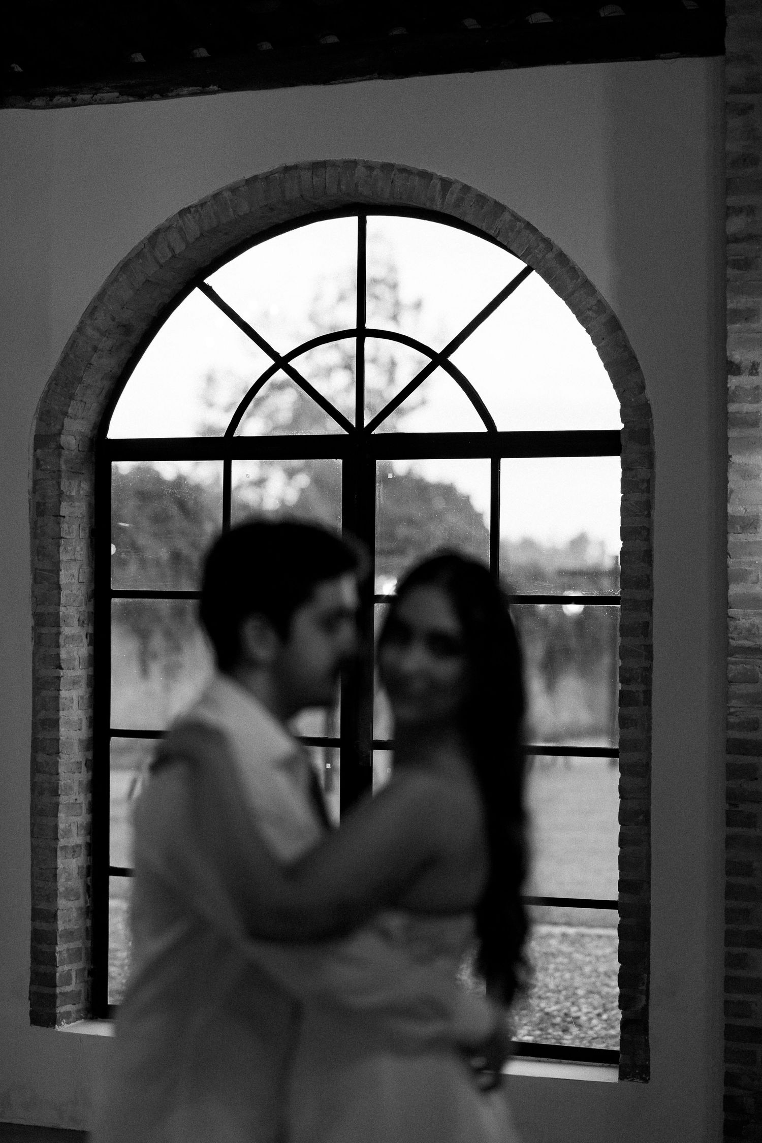 Black and white photo of a couple embracing in front of an arched window, romantic wedding portrait.