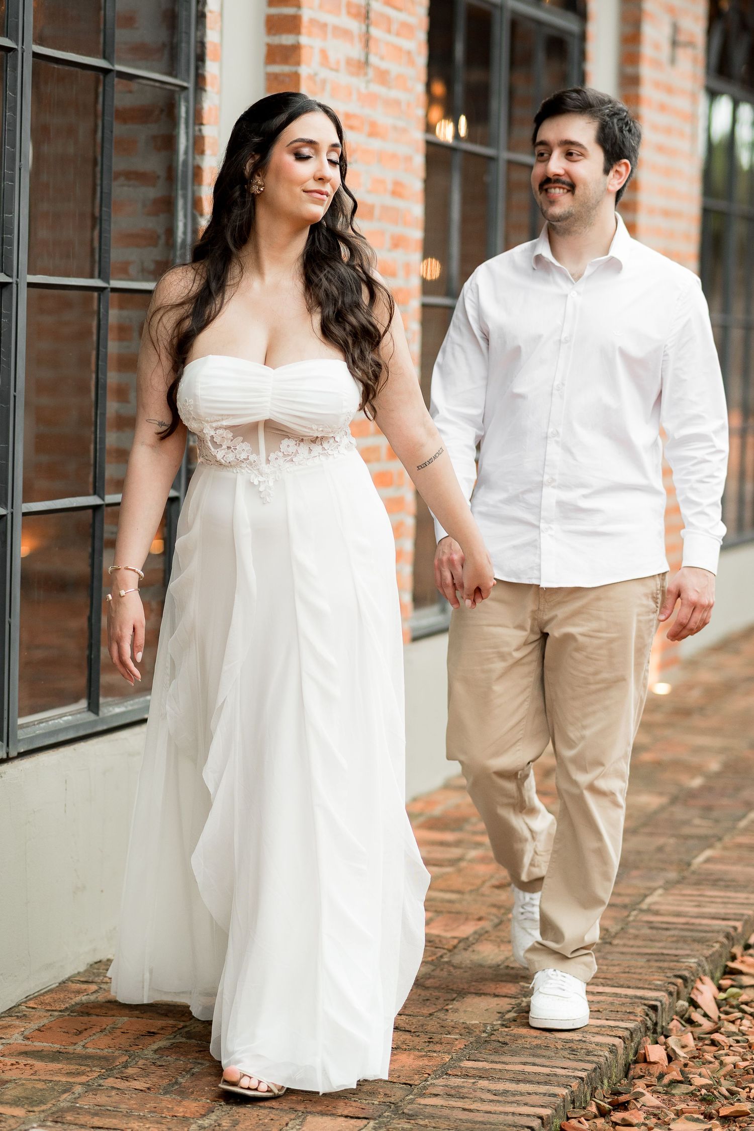Bride in white strapless gown holds hands with groom in white shirt and khakis walking on brick path.