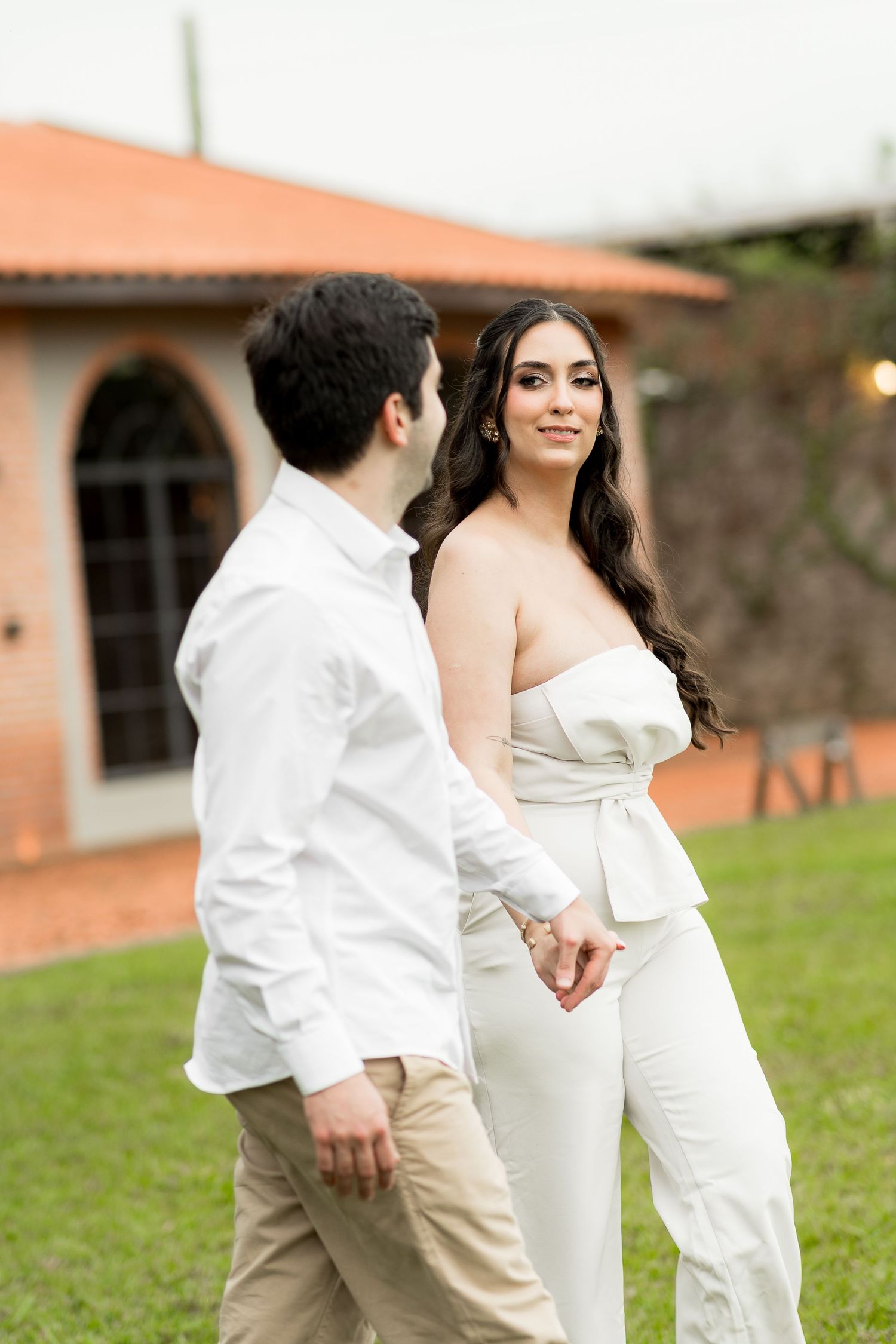 Elegant couple holding hands outdoors, woman in white strapless jumpsuit with bow, man in white shirt and khaki pants.
