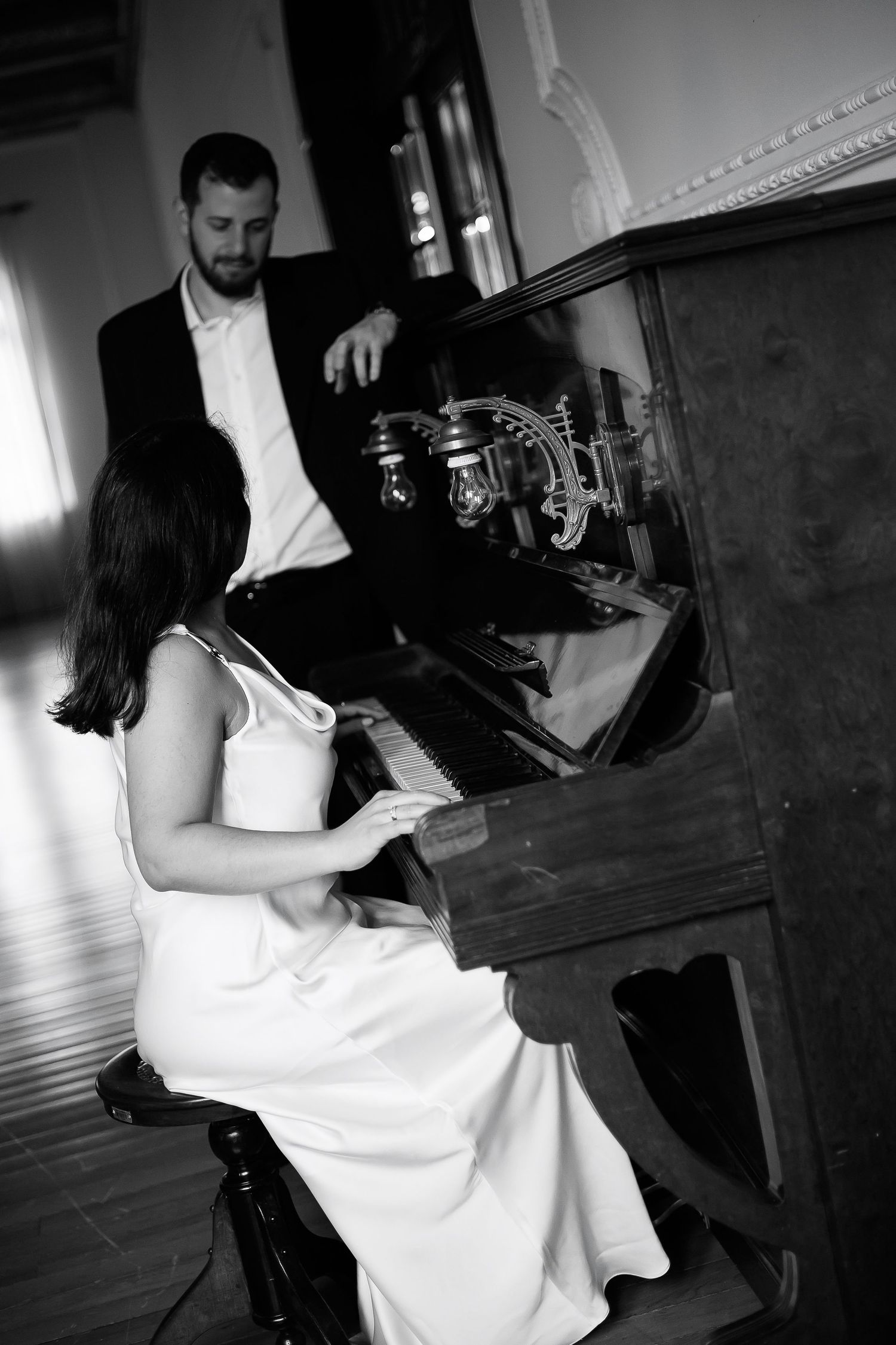 Black and white photo of bride playing upright piano with groom standing behind her in formal attire.