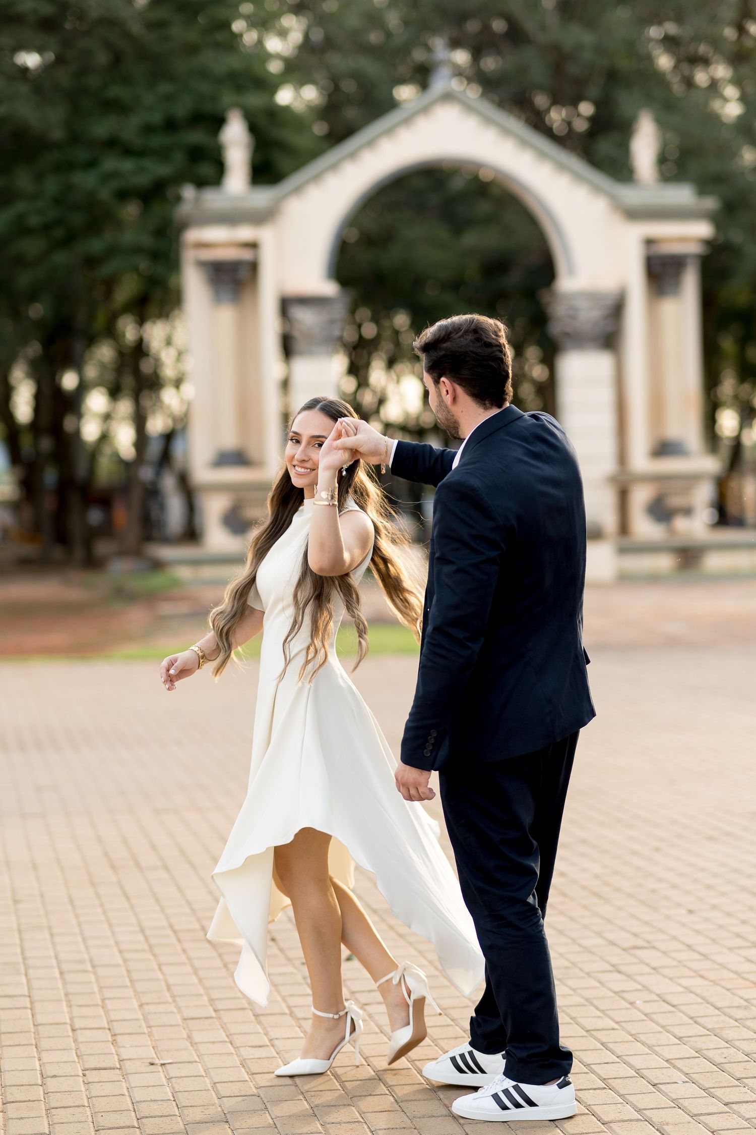 Bride in white dress being twirled by groom in dark suit on brick plaza with ornate arch backdrop.