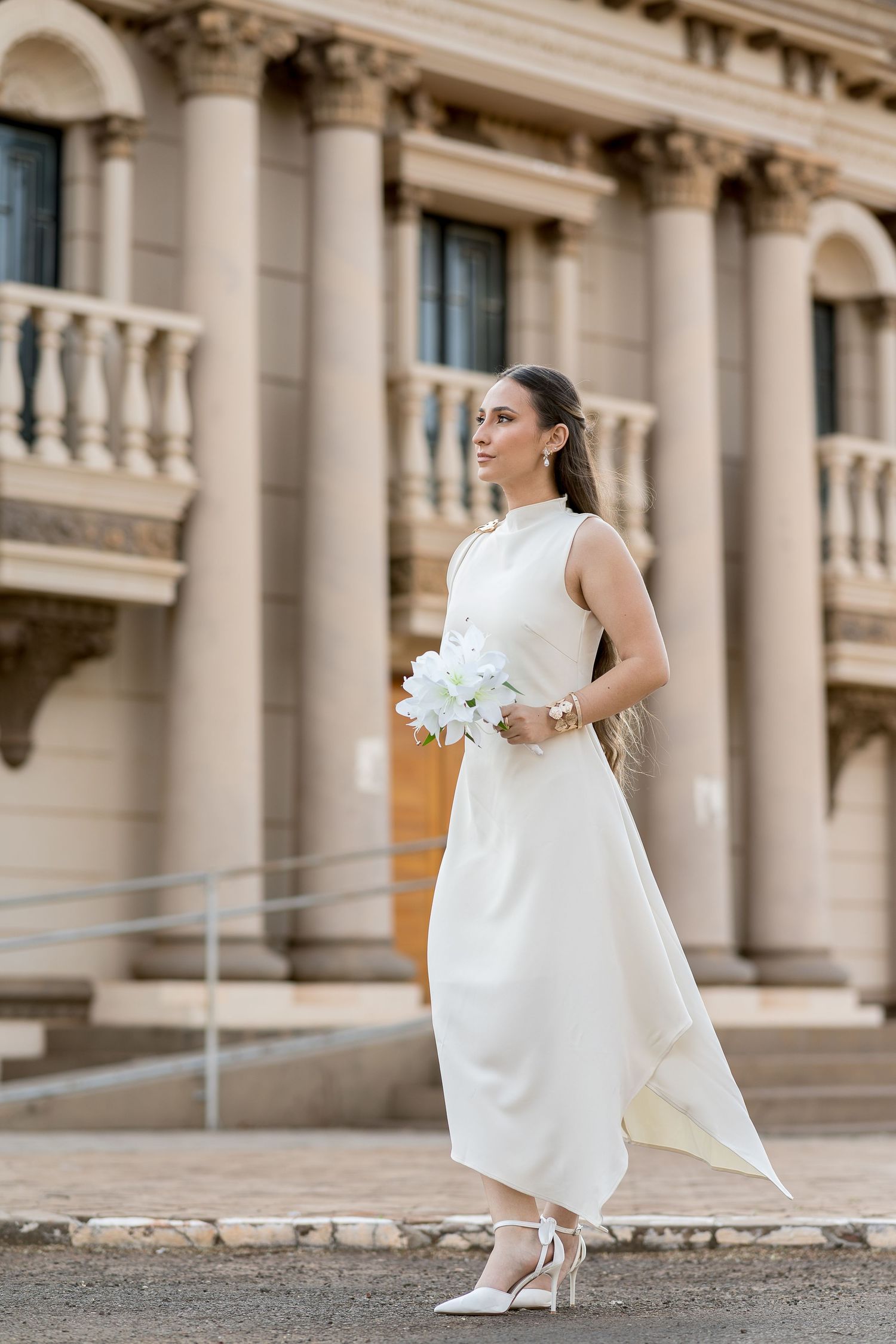 Elegant bride in white midi dress holding bouquet, posing before classical stone building with columns.