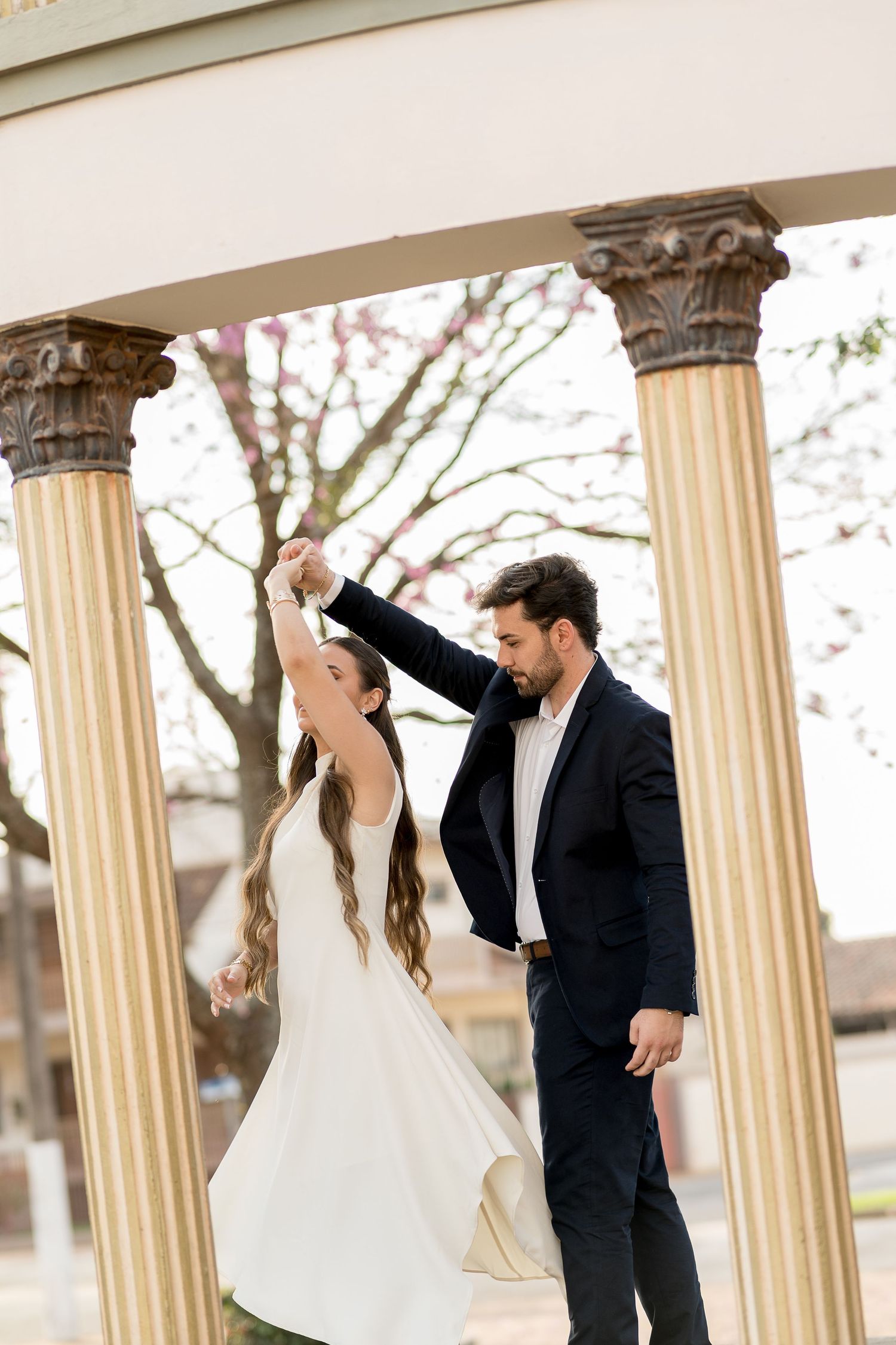 A groom twirls his bride under a classical columned gazebo outdoors during their wedding dance.