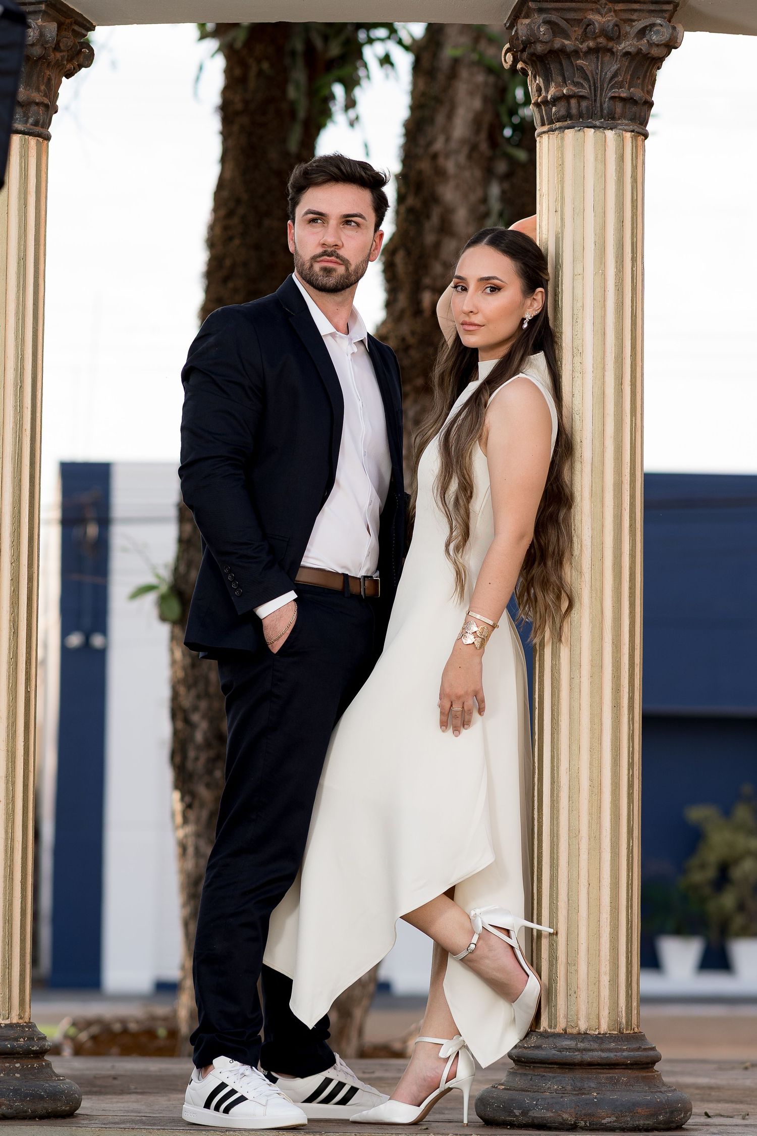 Elegant couple posing between classical columns, man in black suit, woman in white asymmetric dress and heels.
