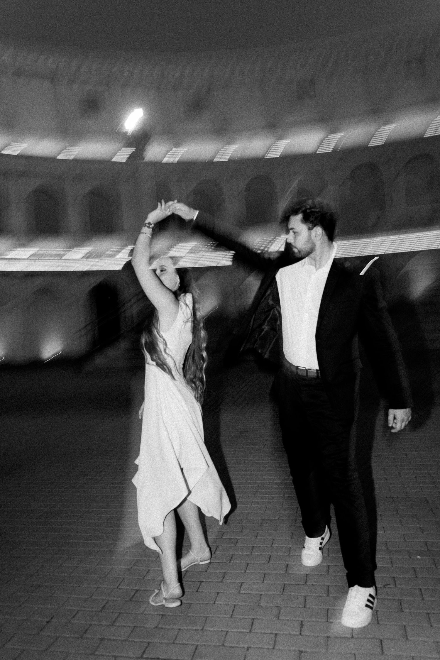 Black and white photo of a couple dancing outdoors at night near an illuminated historic amphitheater.