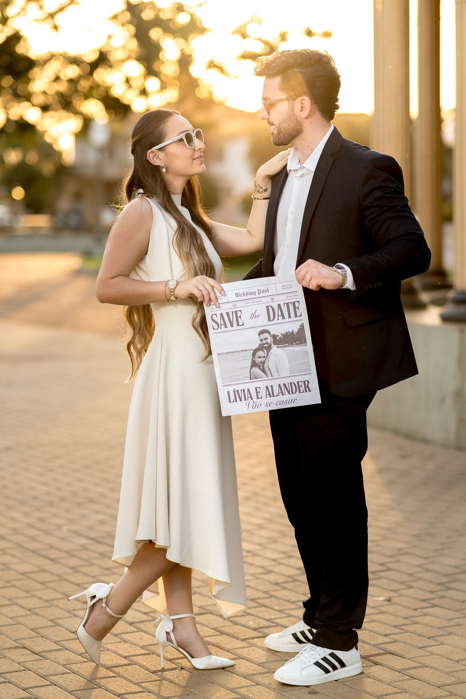 Couple in elegant attire holding a Save the Date newspaper announcement for Livia and Alander at golden hour.
