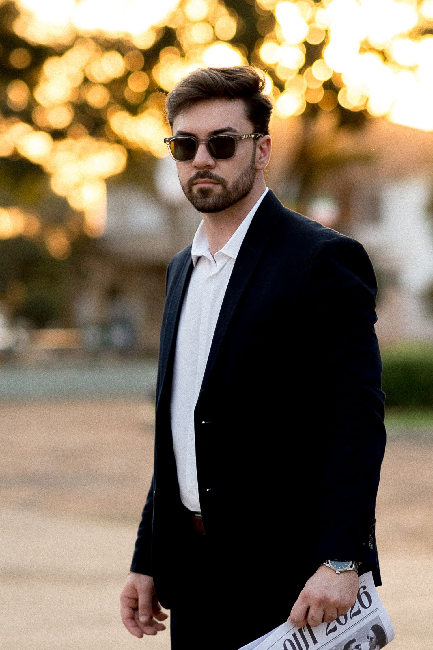Stylish bearded man in black blazer and sunglasses holding newspaper outdoors at golden hour.