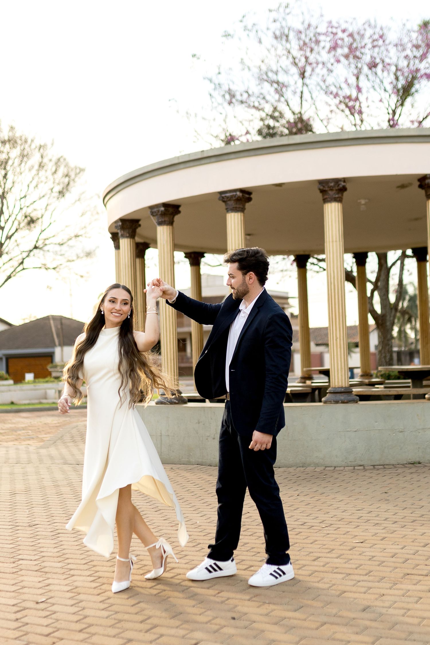 Couple dancing outdoors near a classical rotunda, woman in white dress, man in dark suit, on a brick plaza.