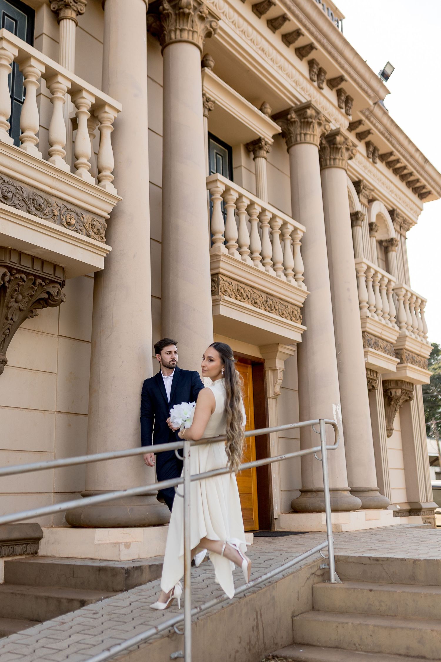 Bride and groom pose on steps of elegant neoclassical building with tall columns, bride holding white bouquet in wedding attire.