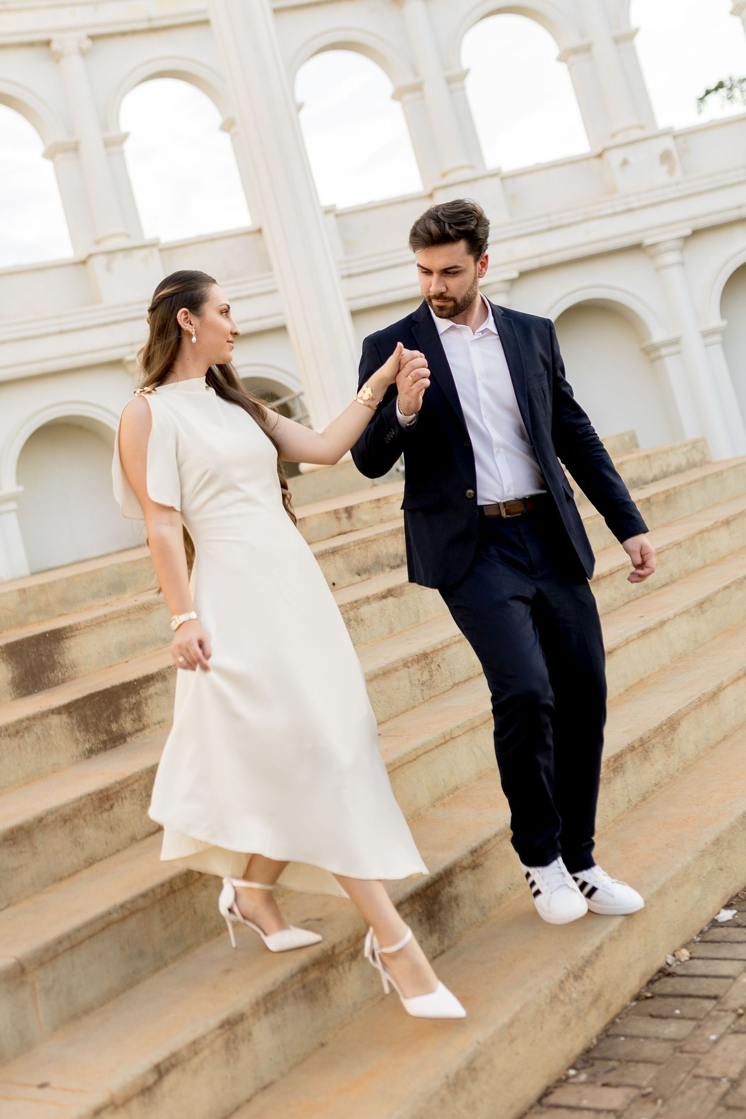 Elegant couple holding hands on grand staircase, woman in white dress and heels, man in navy suit.
