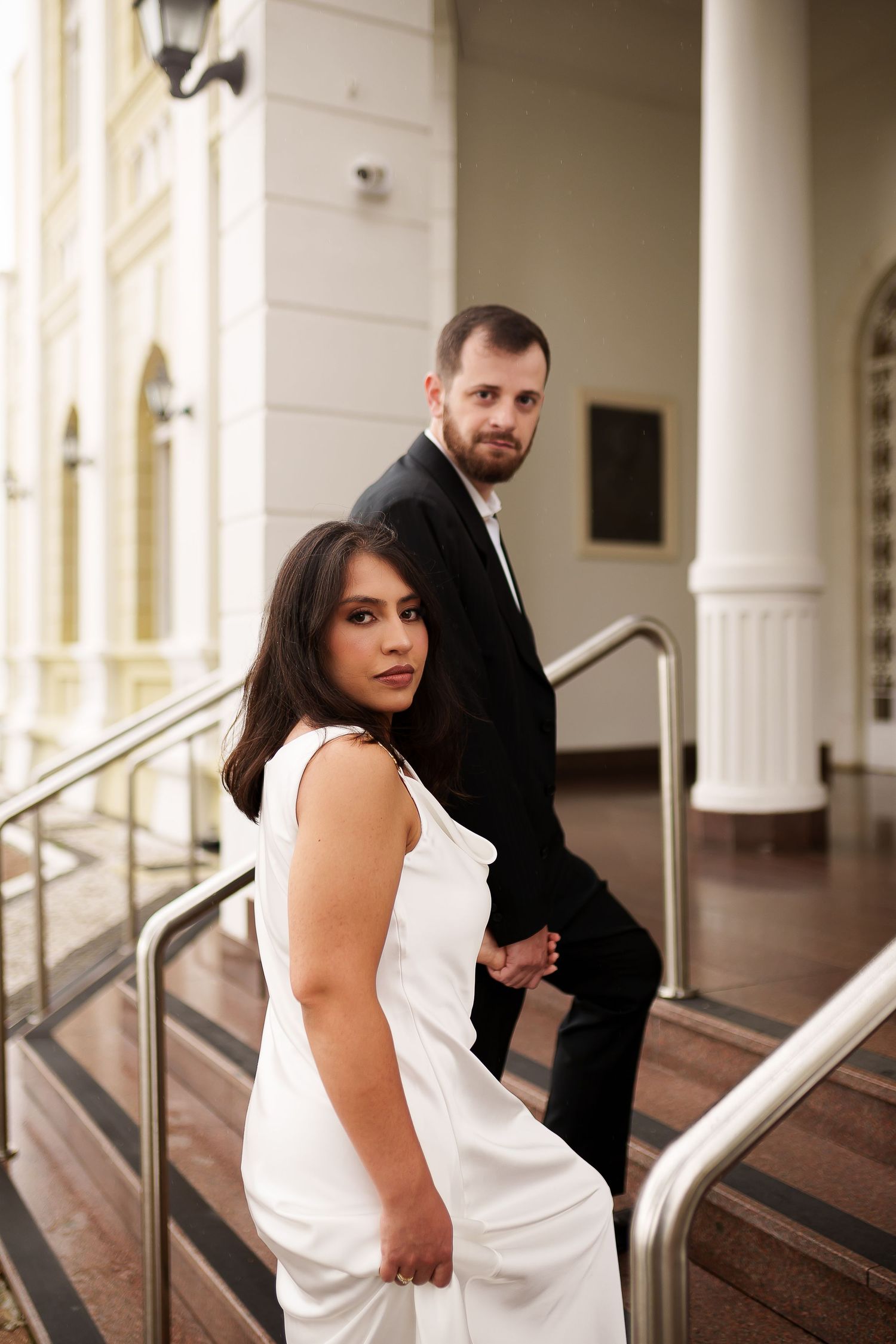 Elegant couple in formal attire posing on marble steps, woman in white gown and man in black suit holding hands.