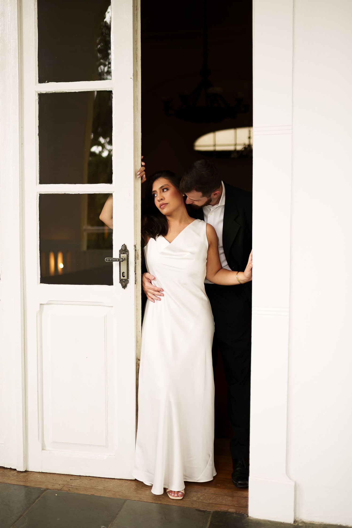 Bride in elegant white satin gown standing in doorway with groom in black suit at wedding venue.