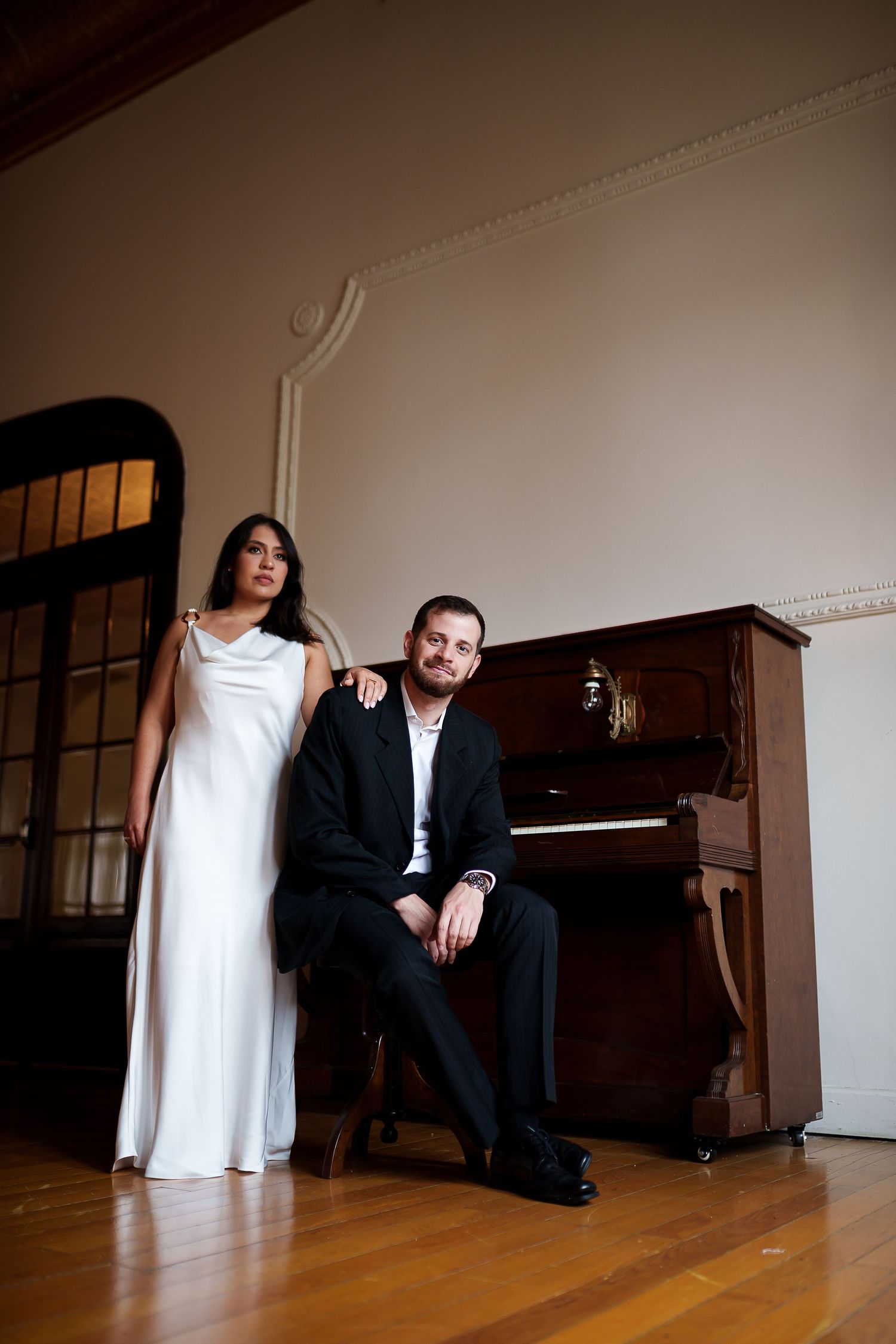 Elegant couple posing by an upright piano, woman in white gown standing beside seated man in black suit.