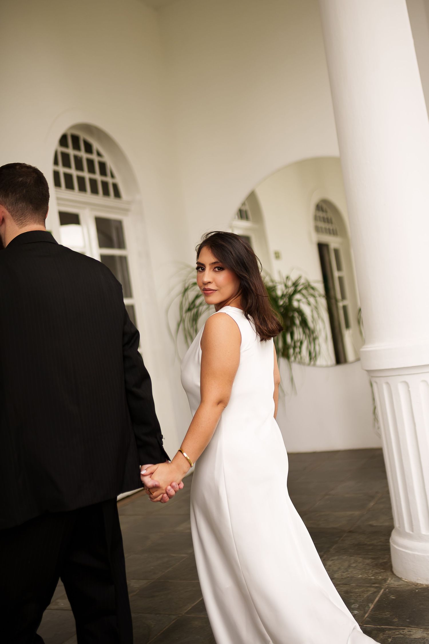 Bride in white gown holds groom's hand, glancing back over shoulder at elegant white columned venue.