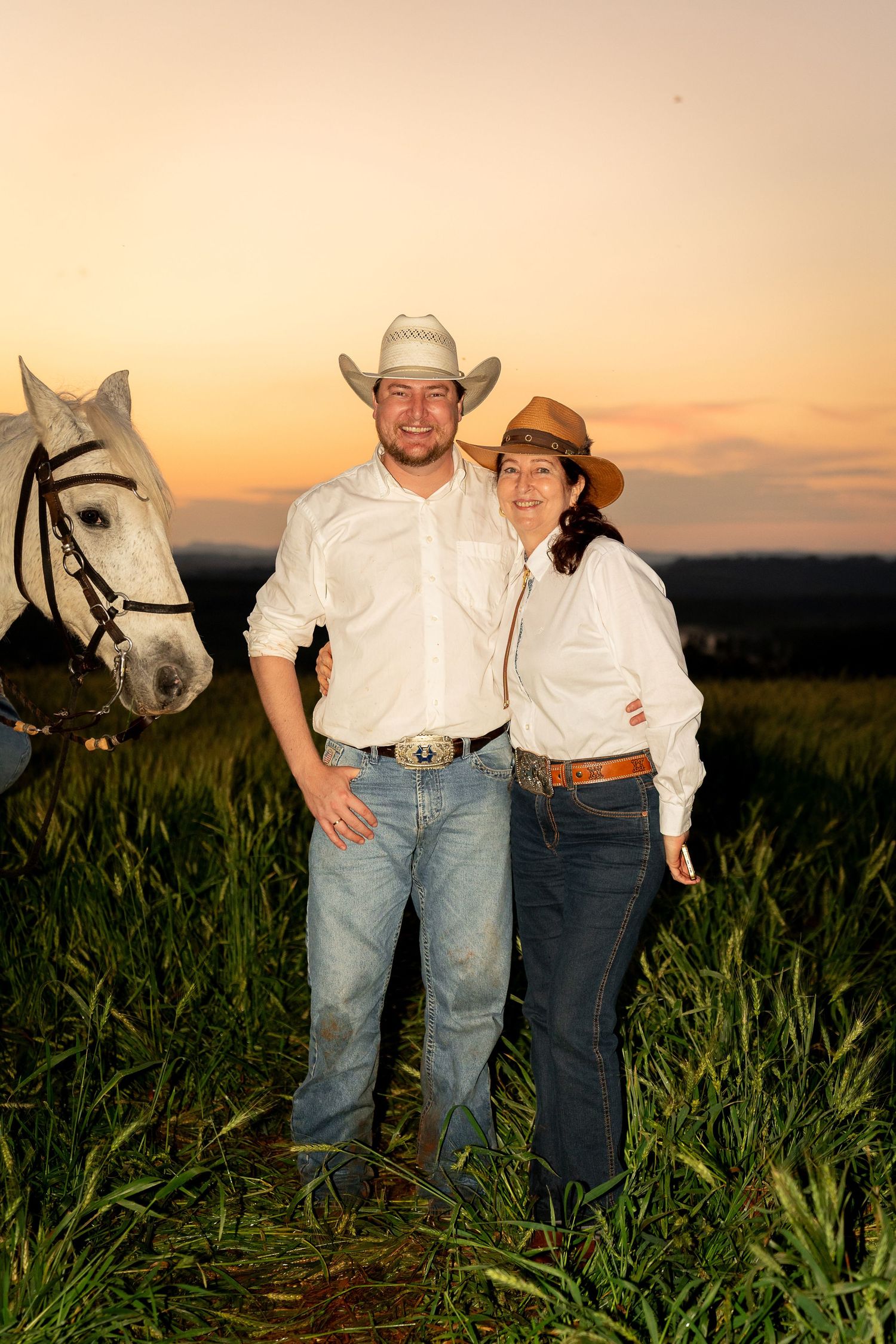 Couple in cowboy hats and white shirts pose at sunset in a green field beside a white horse.