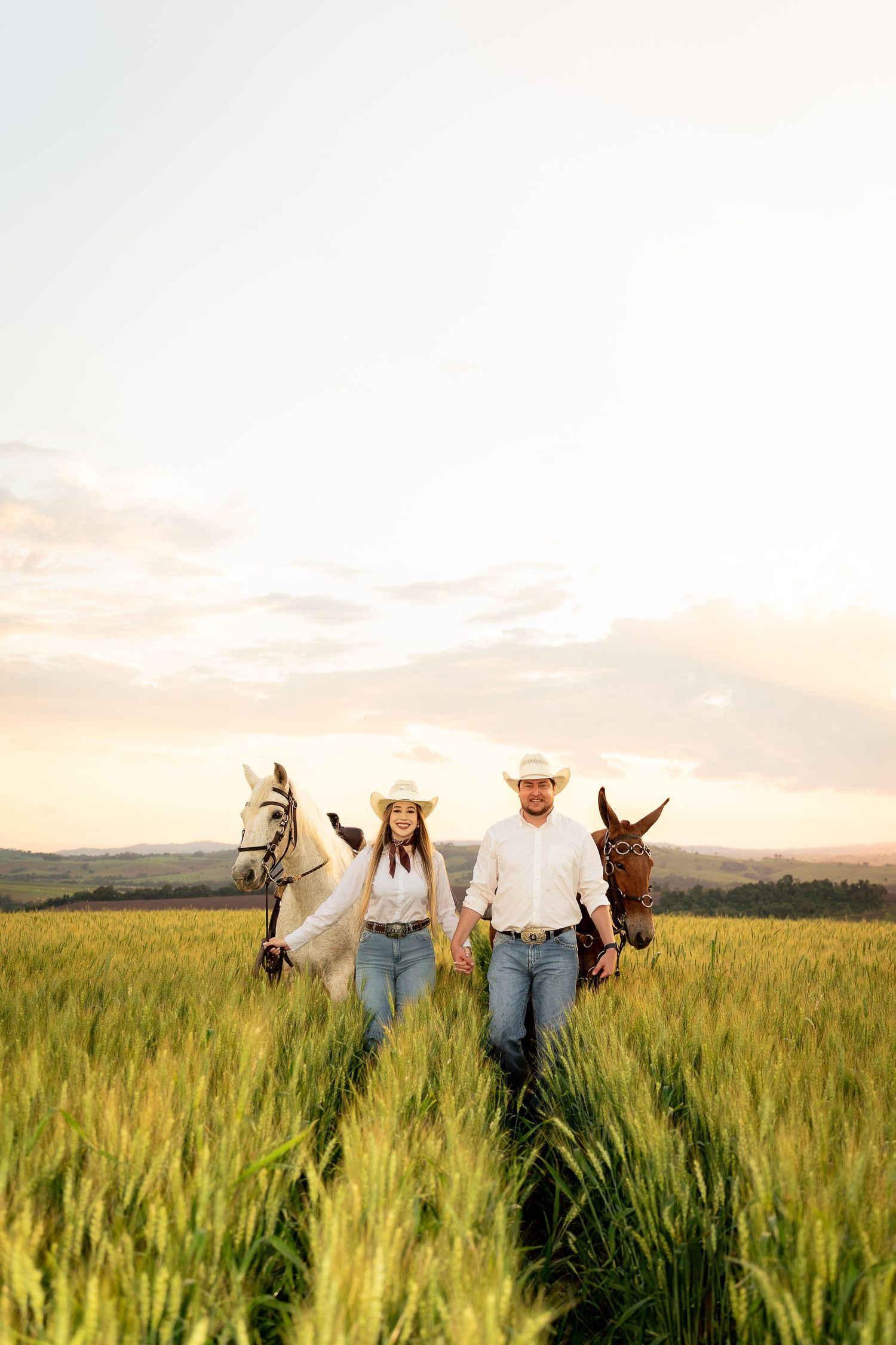 A couple in cowboy hats walk hand-in-hand through a golden wheat field at sunset, each leading a horse.