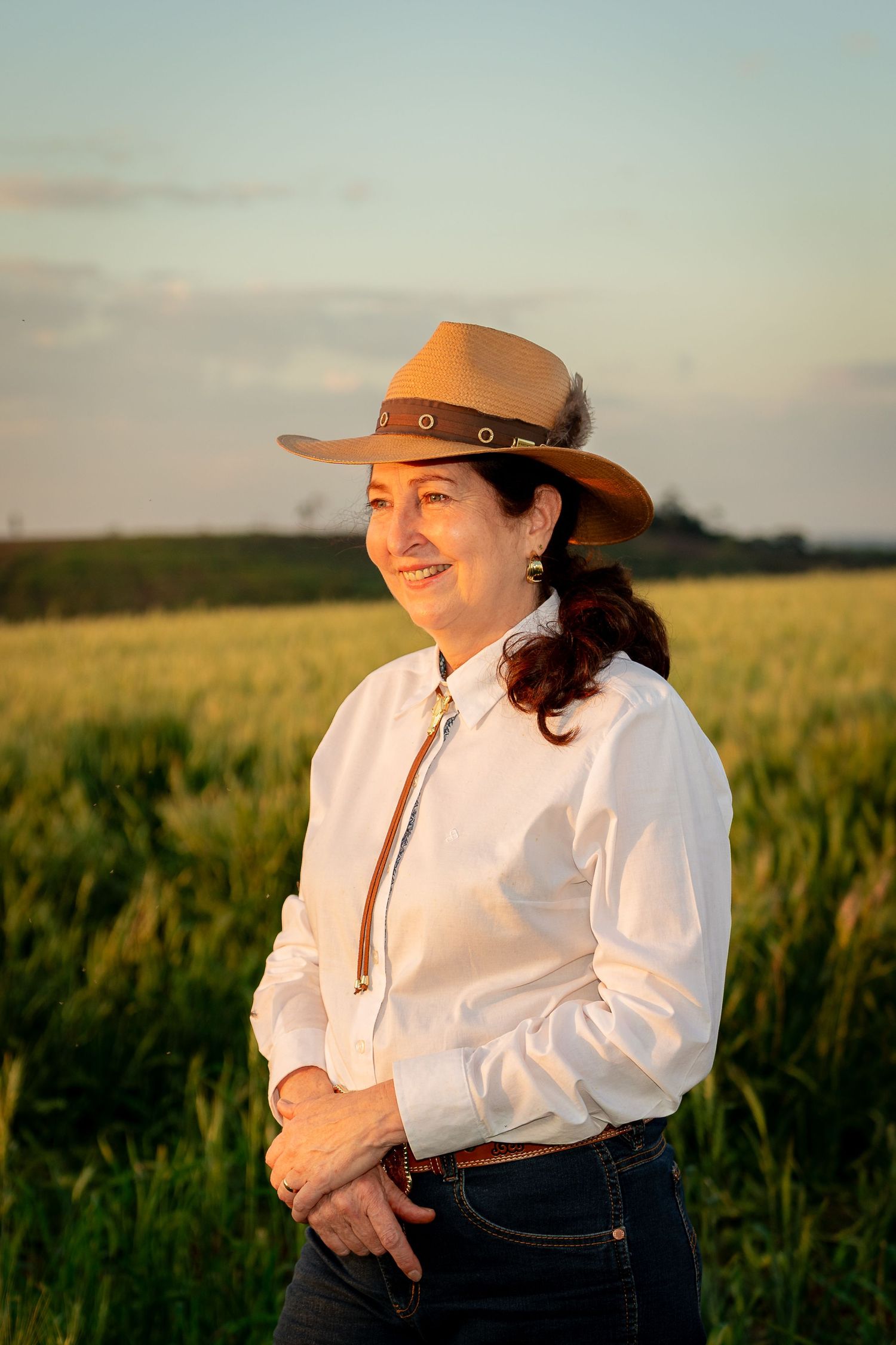 Smiling woman in cowboy hat and white shirt stands in golden field at sunset.