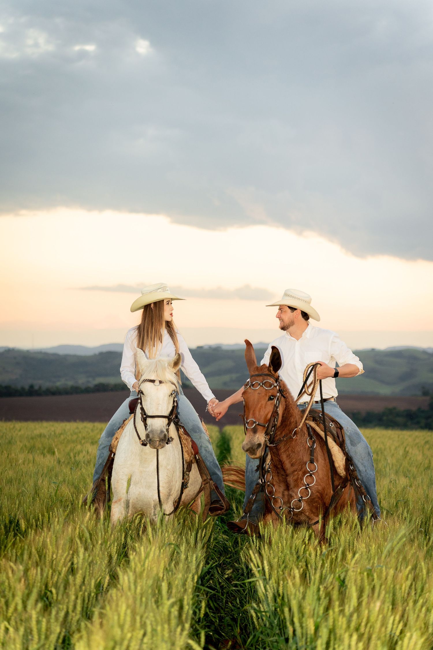 Couple in cowboy hats holding hands while riding horses through a green field at sunset.