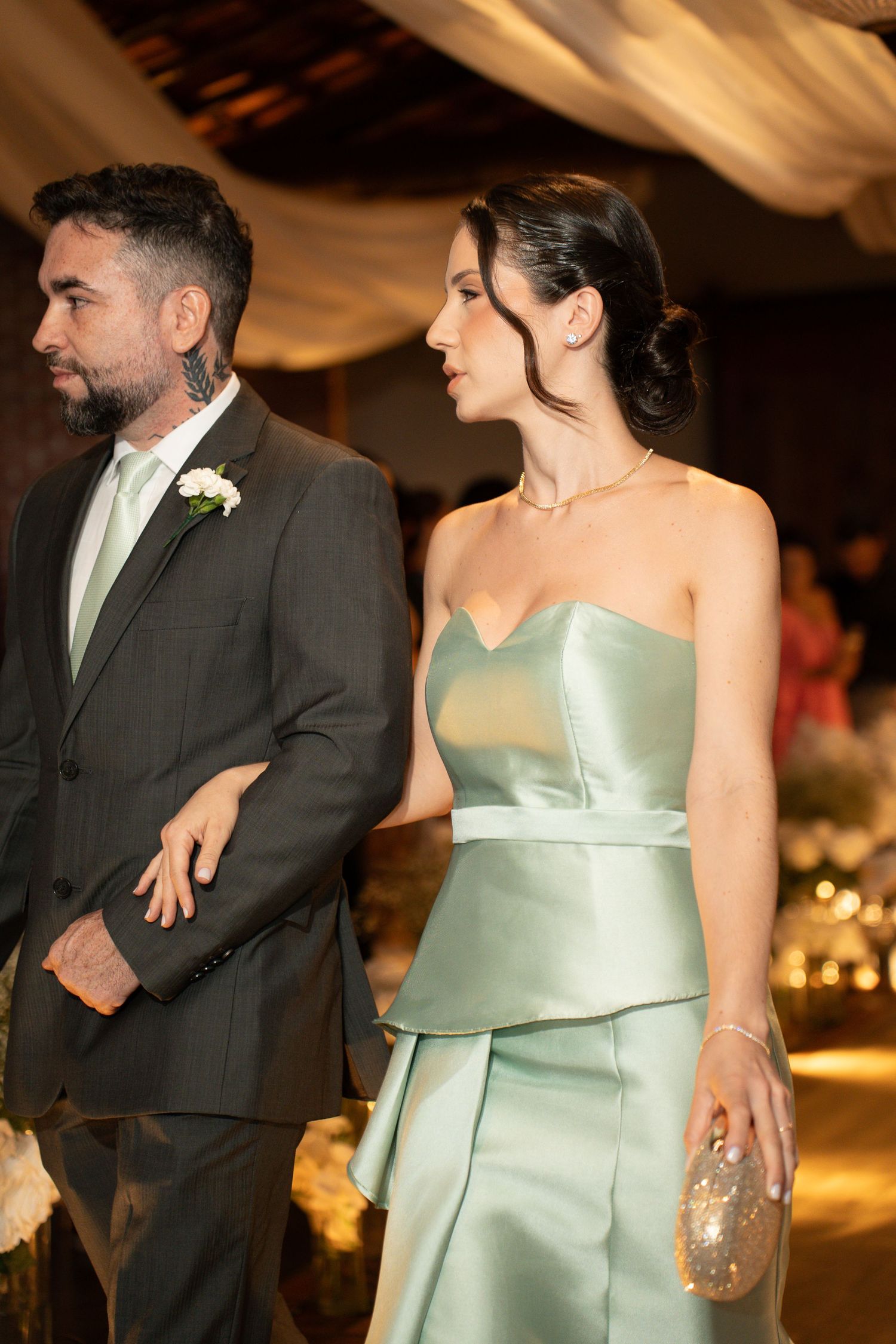 Elegant couple at a wedding, man in gray suit with boutonniere, woman in strapless sage green satin gown holding a gold clutch.
