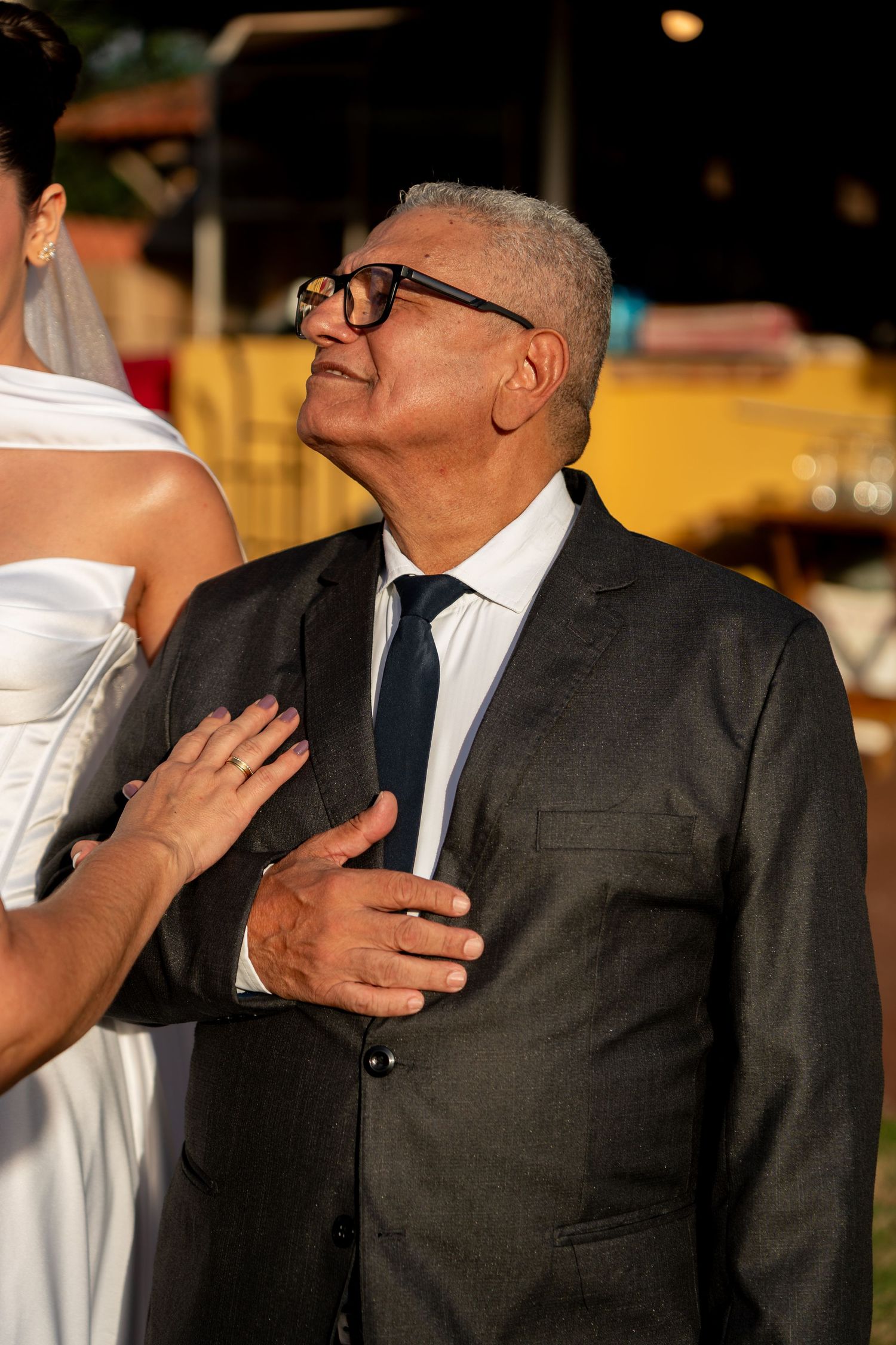 Elderly man in gray suit and glasses at a wedding ceremony, looking up emotionally as bride places hand on his chest.