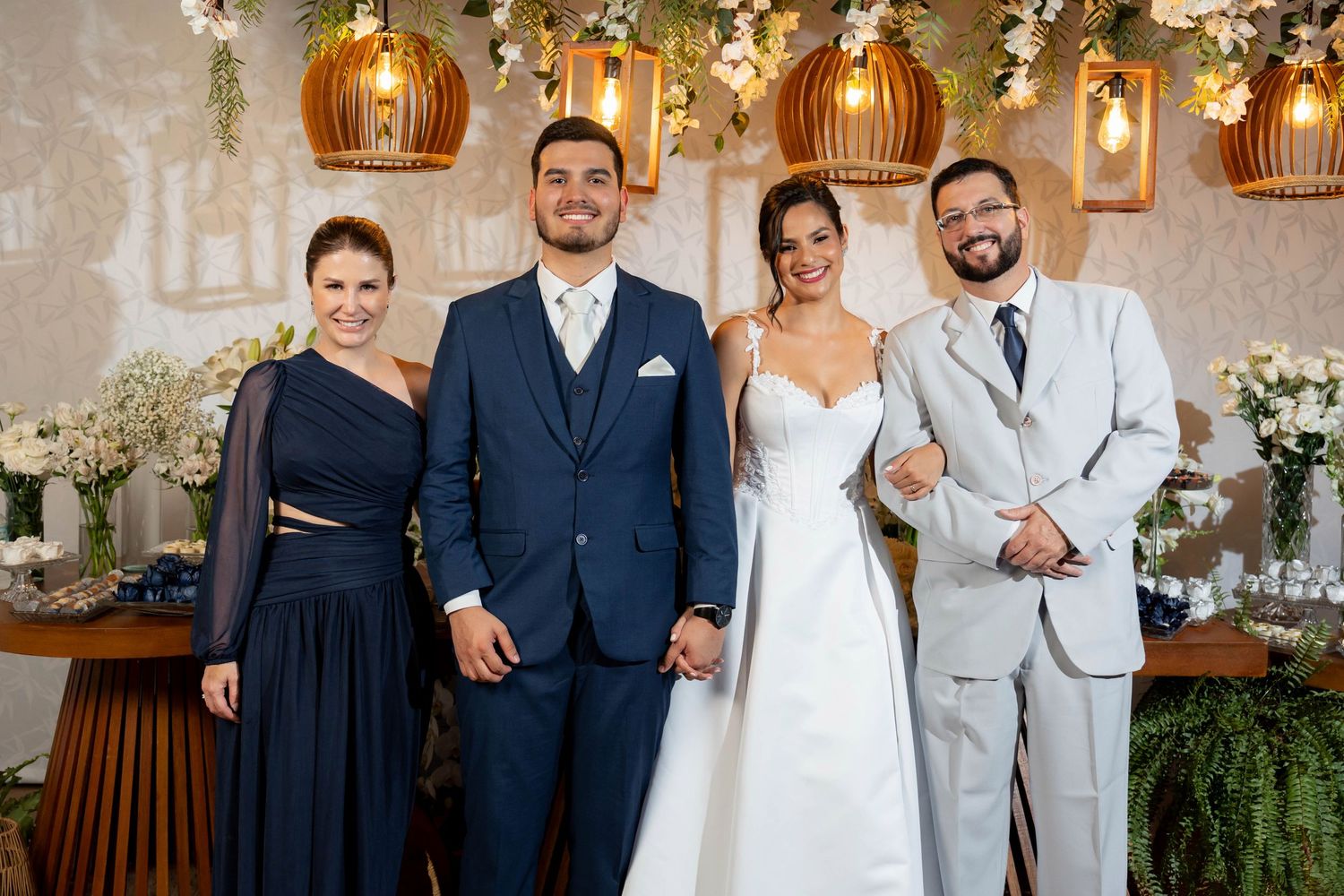 Wedding couple in white dress and navy suit pose with two guests under floral ceiling decor at elegant reception venue.